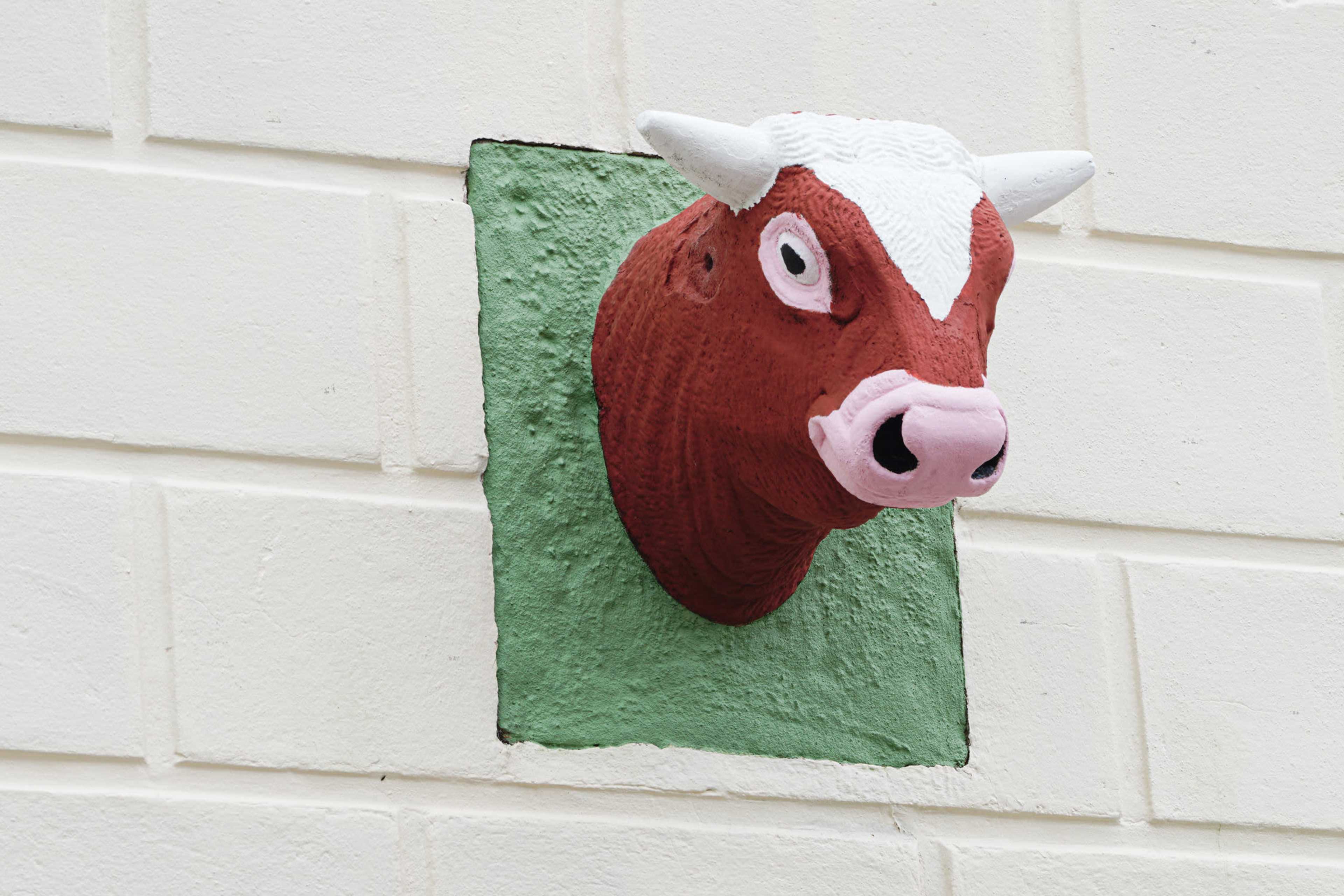 A painted cow's head sculpture protrudes from a green square on a white brick wall.