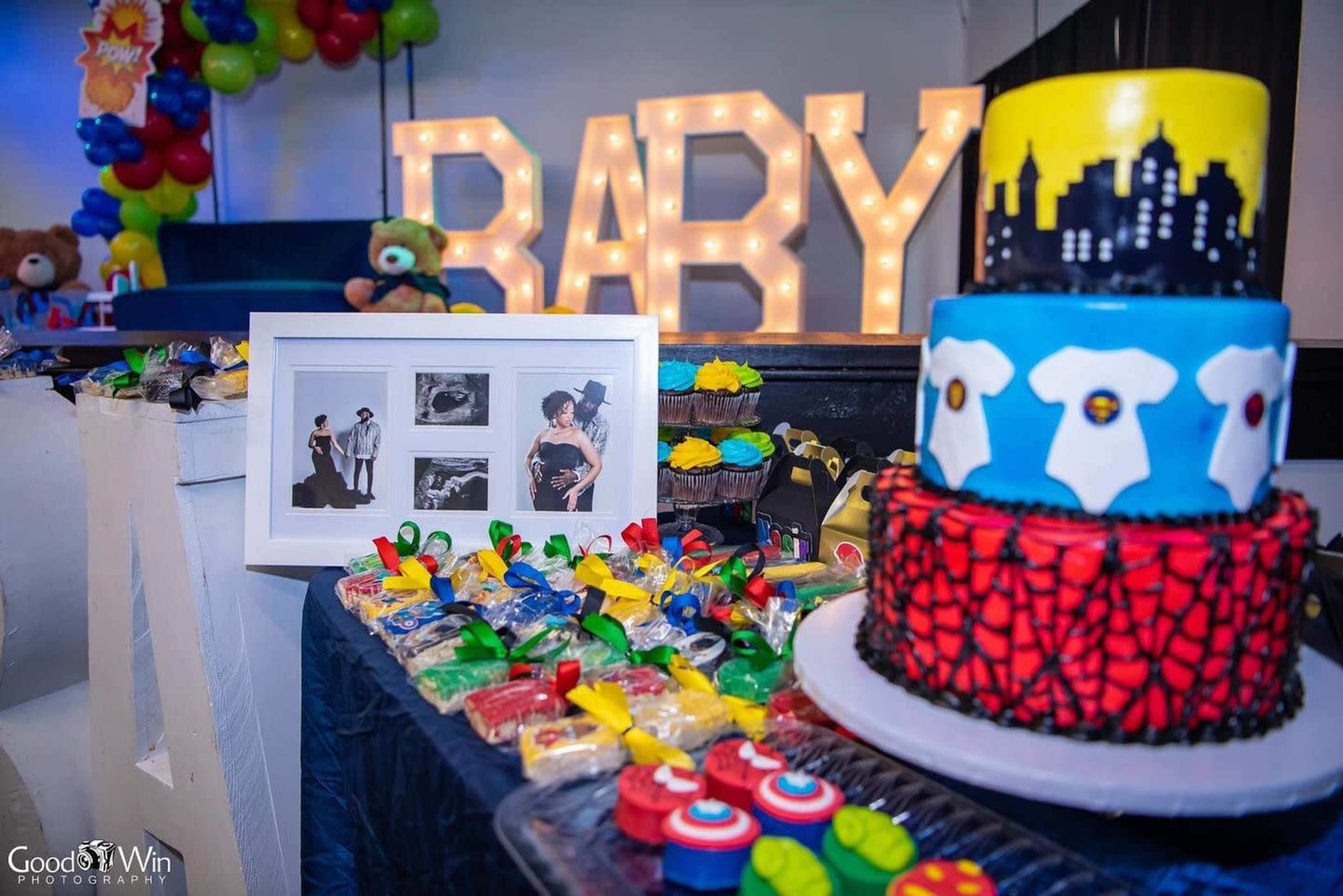 A colorful baby-themed celebration features a decorated cake, treats, and a photo display, with large illuminated letters spelling "BABY" in the background.