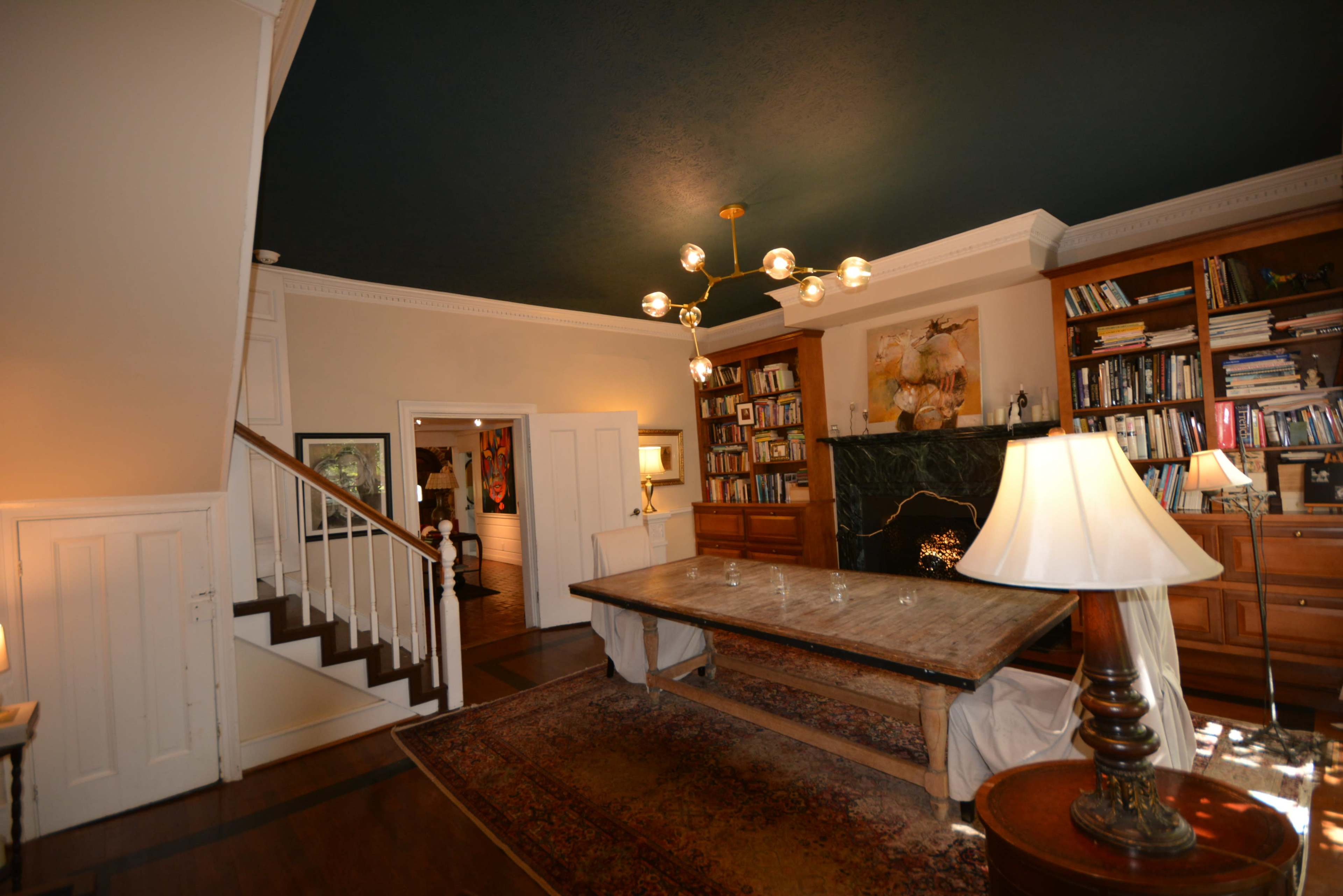 The image shows a spacious living room with a large wooden table, bookshelves filled with books, and a staircase leading to an upper level, all under a dark ceiling.