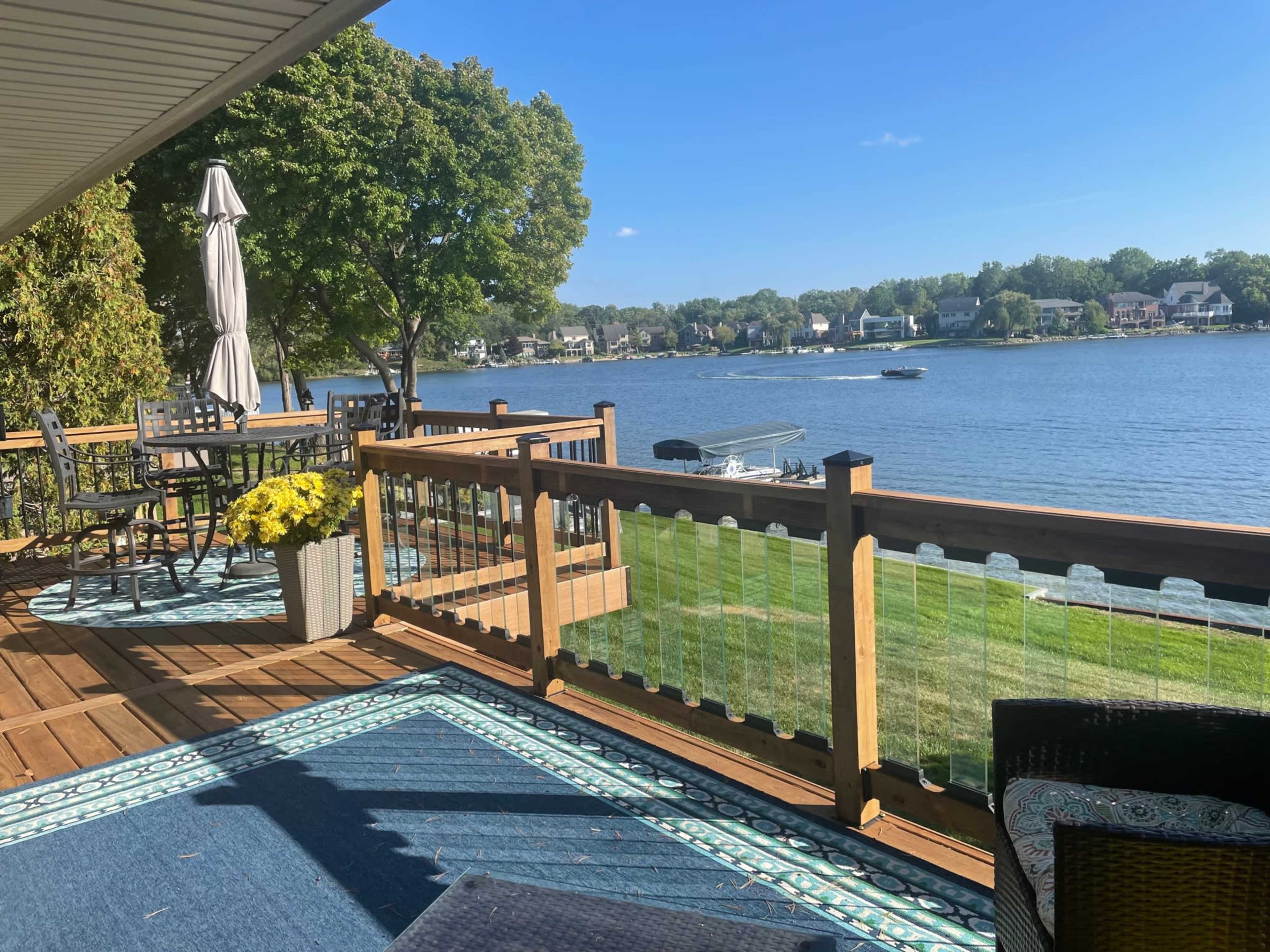 A wooden deck overlooks a lake, featuring a patio table and chairs, an umbrella, and a boat passing by on the water.