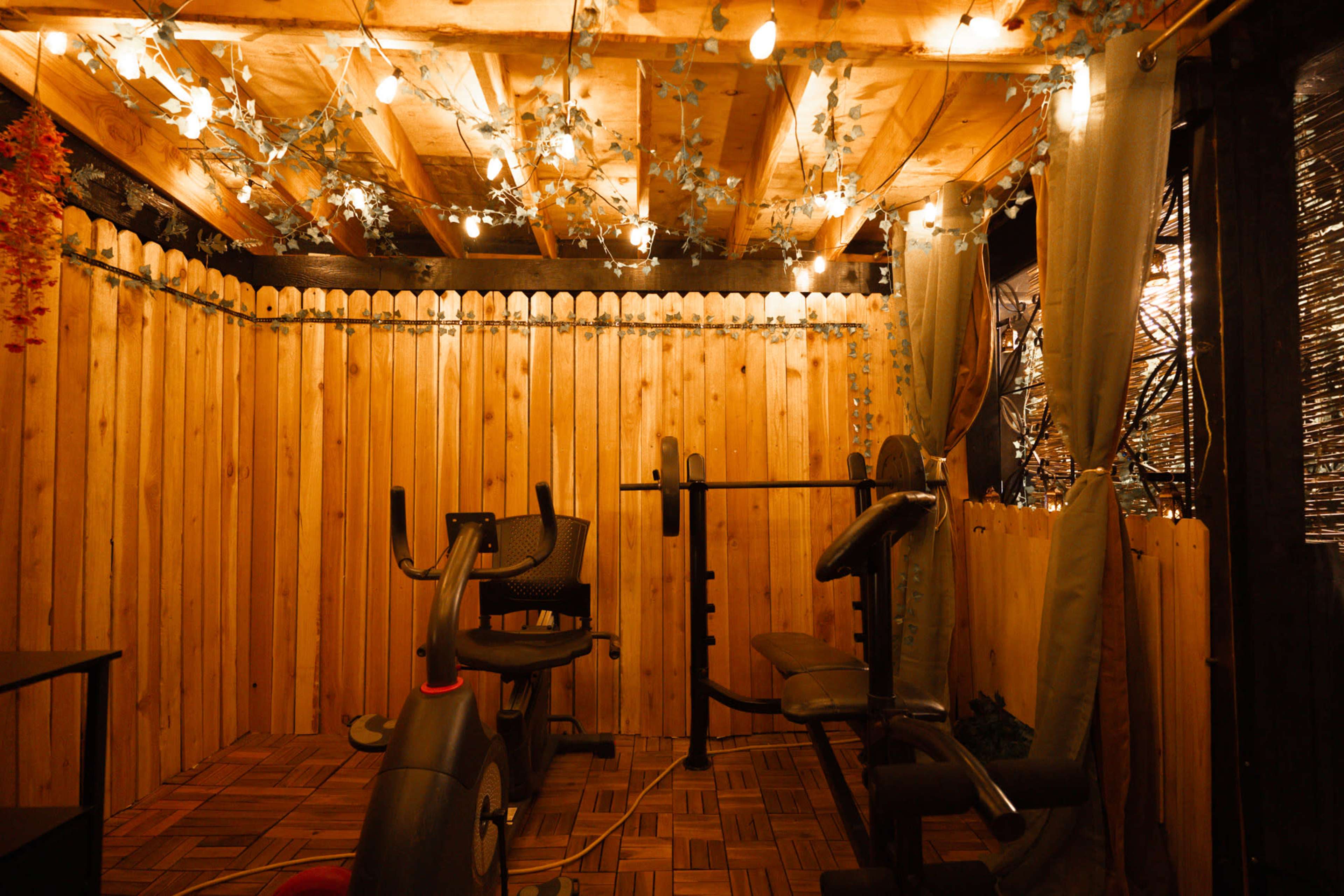 The scene features a cozy home gym with wooden walls, exercise equipment, and soft lighting from string lights overhead.