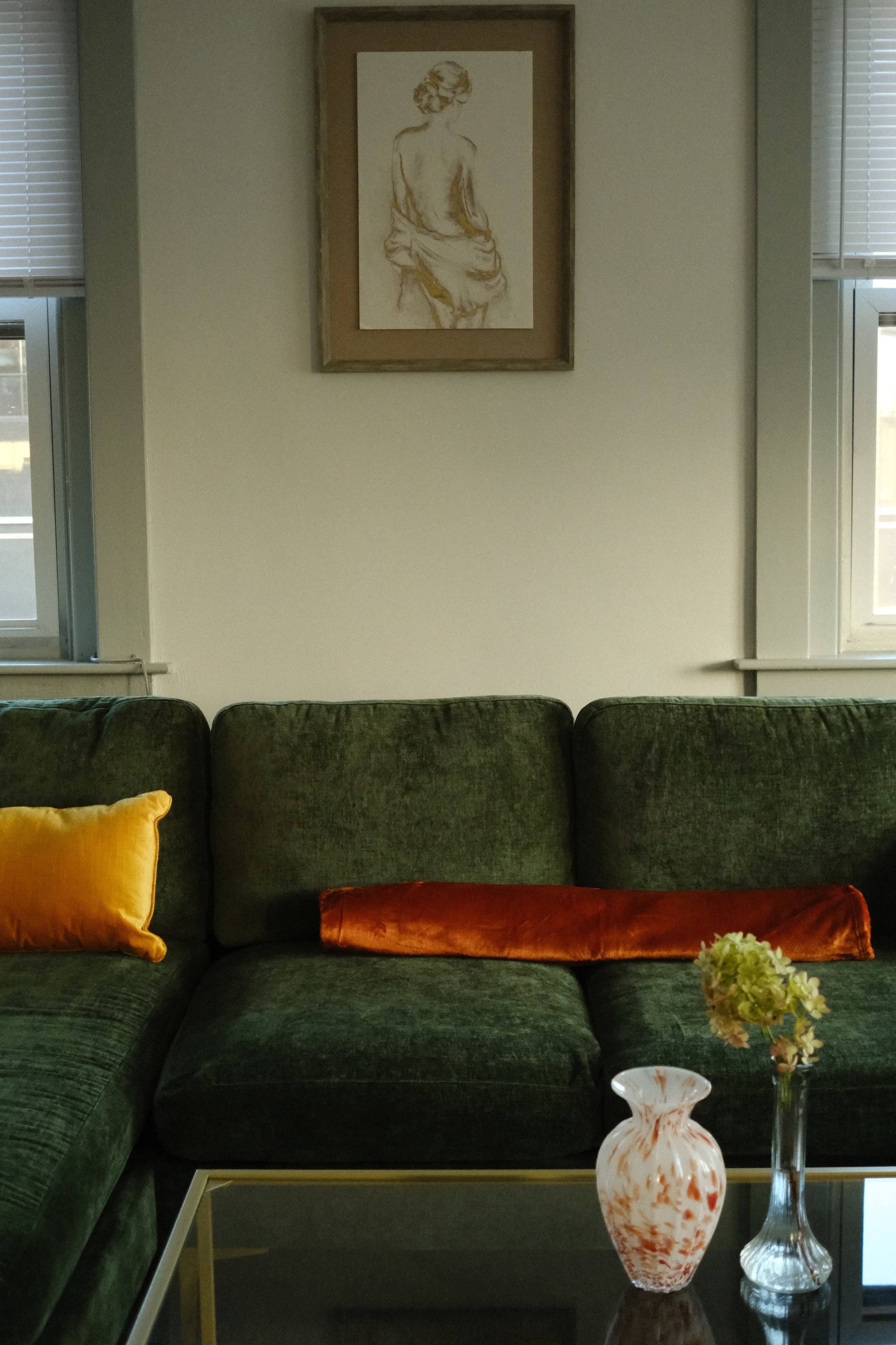 A green couch with orange and gold cushions is positioned in front of a wall featuring a framed artwork, accompanied by a glass table with two vases and a flower.
