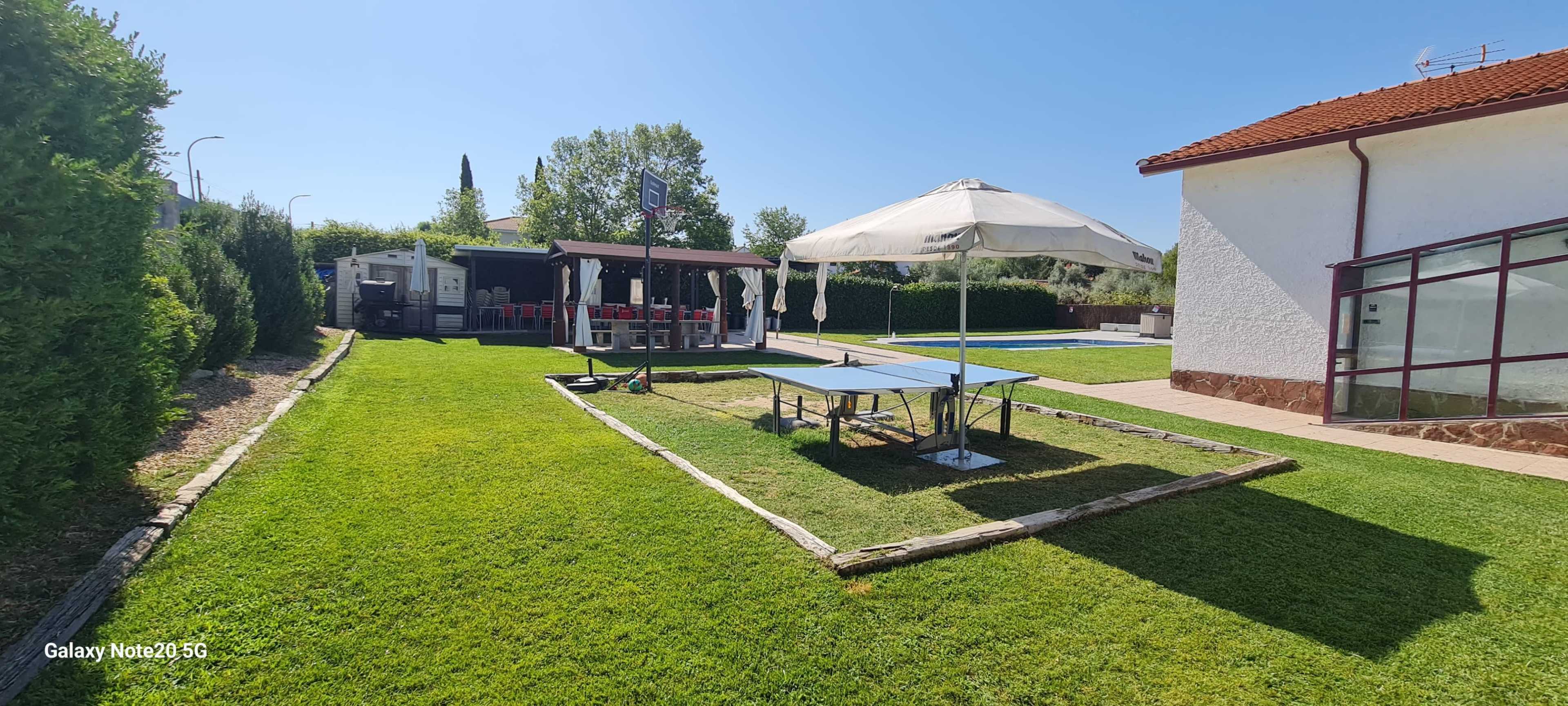 The image shows a backyard area with a ping pong table, a shaded seating area, and a swimming pool in the background, surrounded by well-maintained grass and trees.