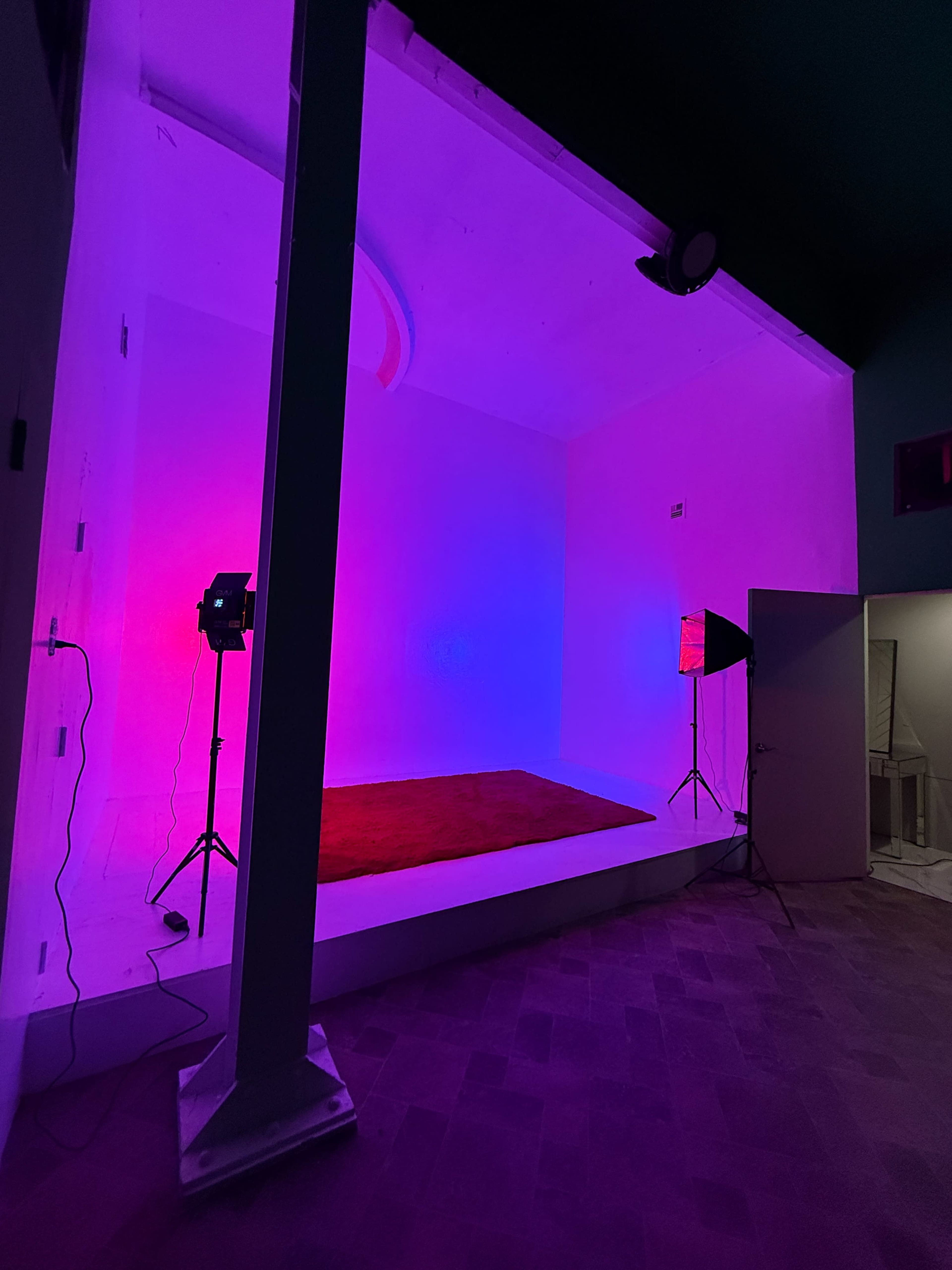 The image shows an empty room with a raised platform, illuminated in pink and purple lighting, and equipped with photography lights on stands.