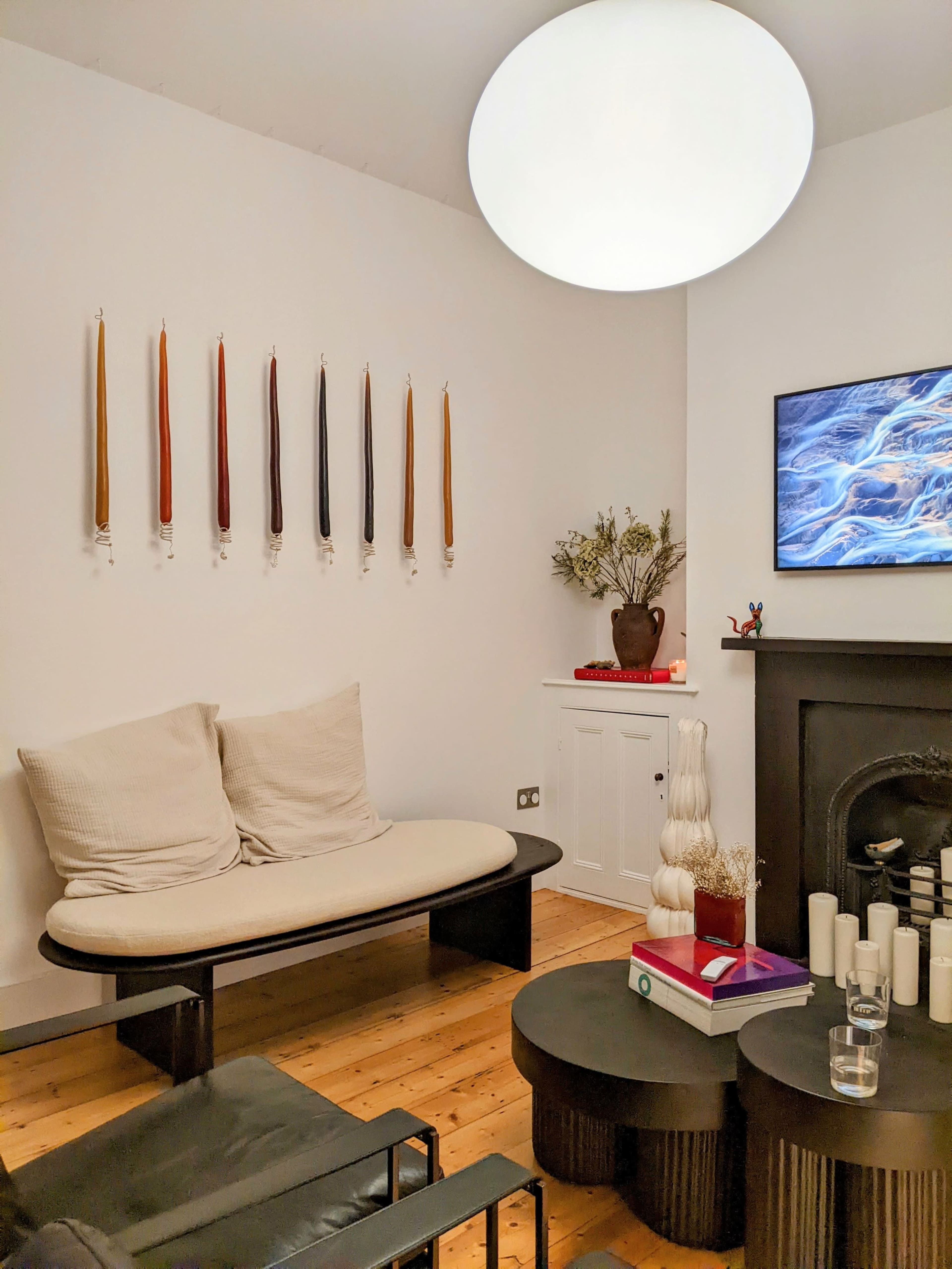 The image depicts a minimalist living room with a light-colored sofa, a dark coffee table, candles on display, and a wall of colorful taper candles.