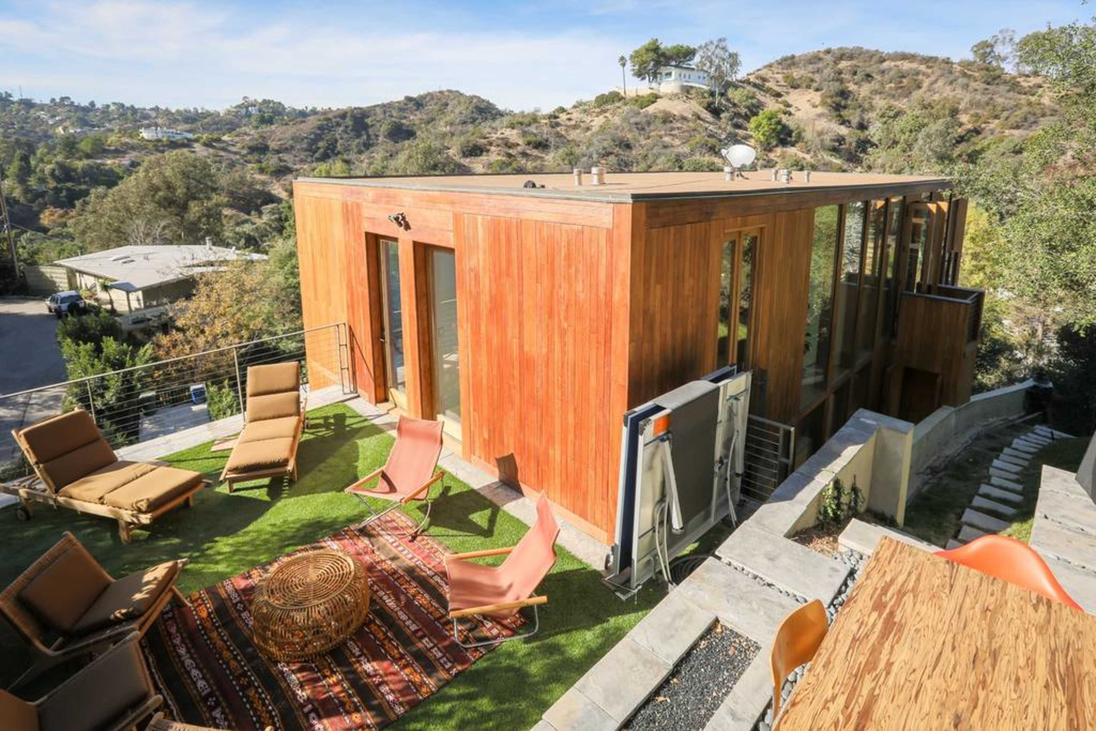A modern wooden house is situated on a hillside, surrounded by outdoor seating and a patterned rug.