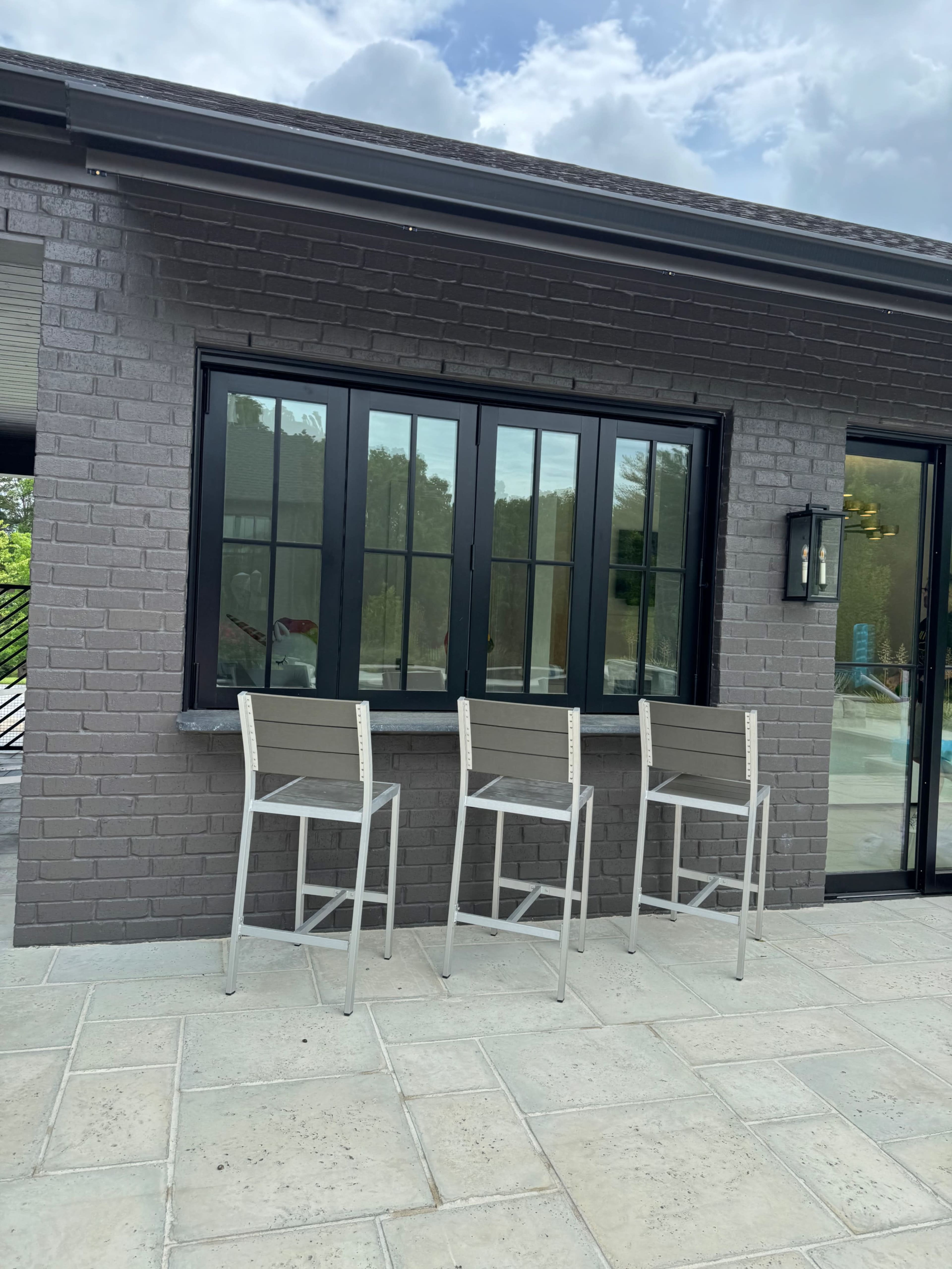 Three tall outdoor bar stools lined up against a window of a gray-brick building, with a view of greenery in the background.