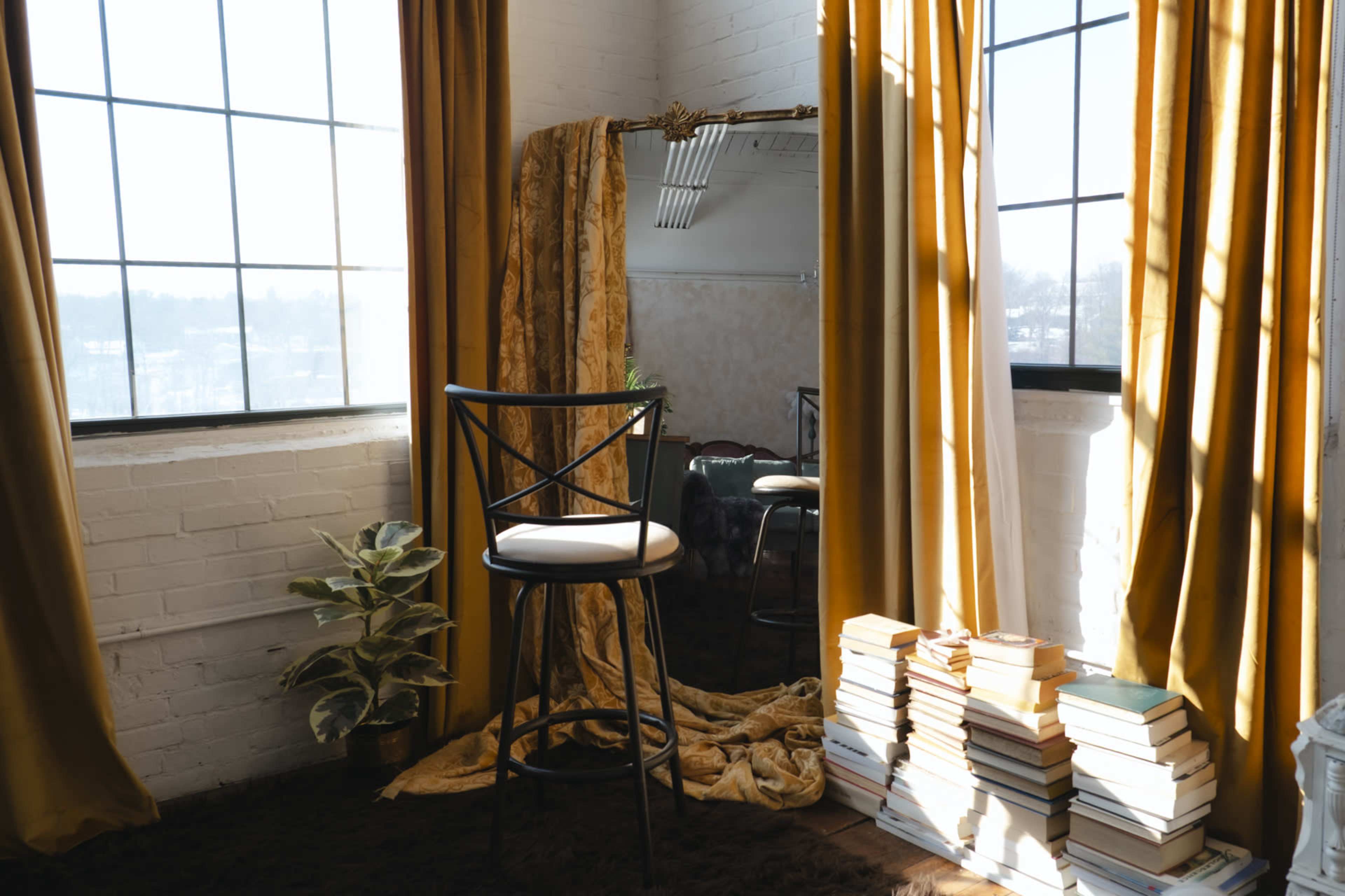 A tall bar stool sits beside a stack of books in a sunlit room with large windows and golden curtains.