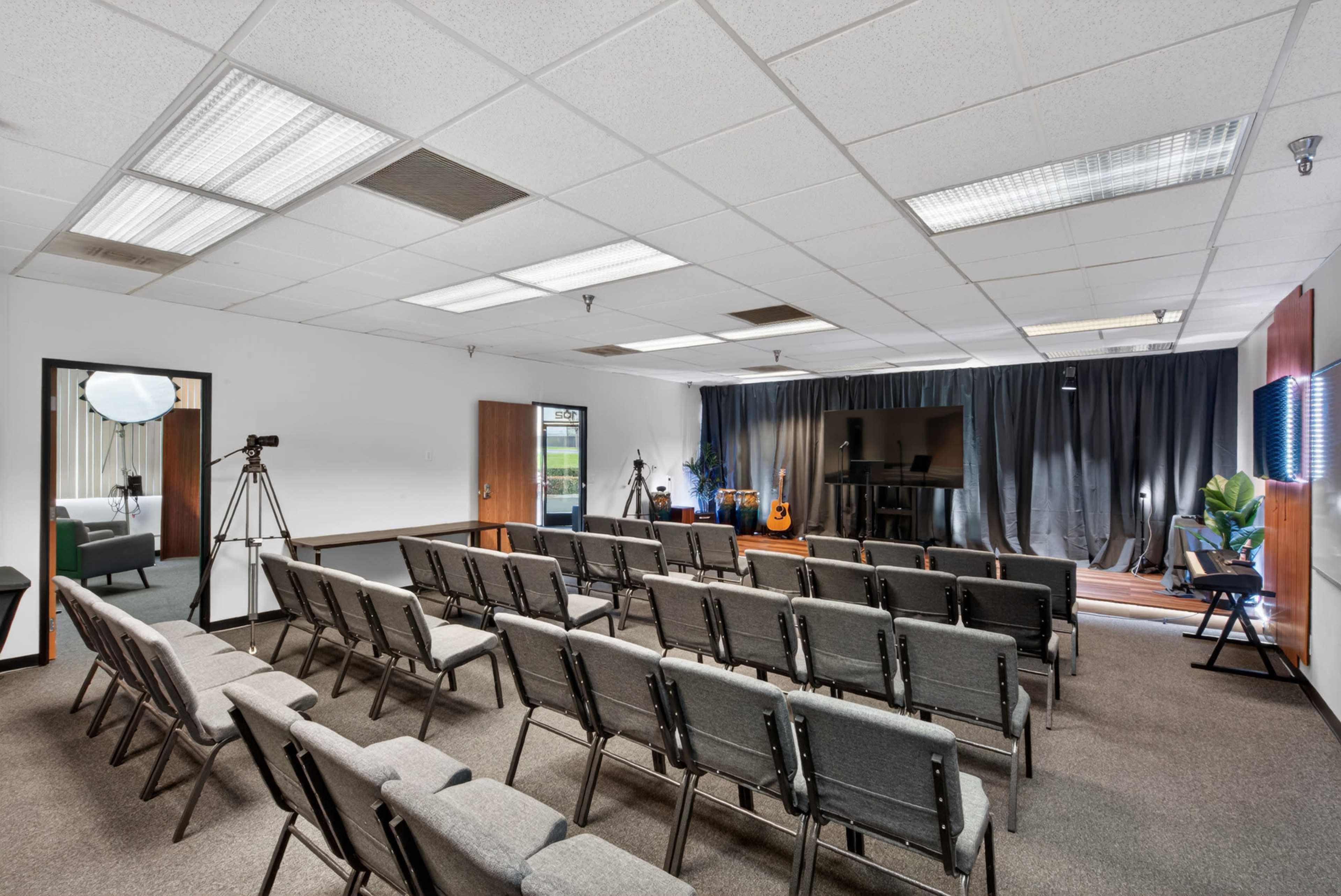 The image shows a spacious room arranged with rows of gray folding chairs facing a black curtain backdrop, with a few cameras and equipment set up for a presentation or recording.