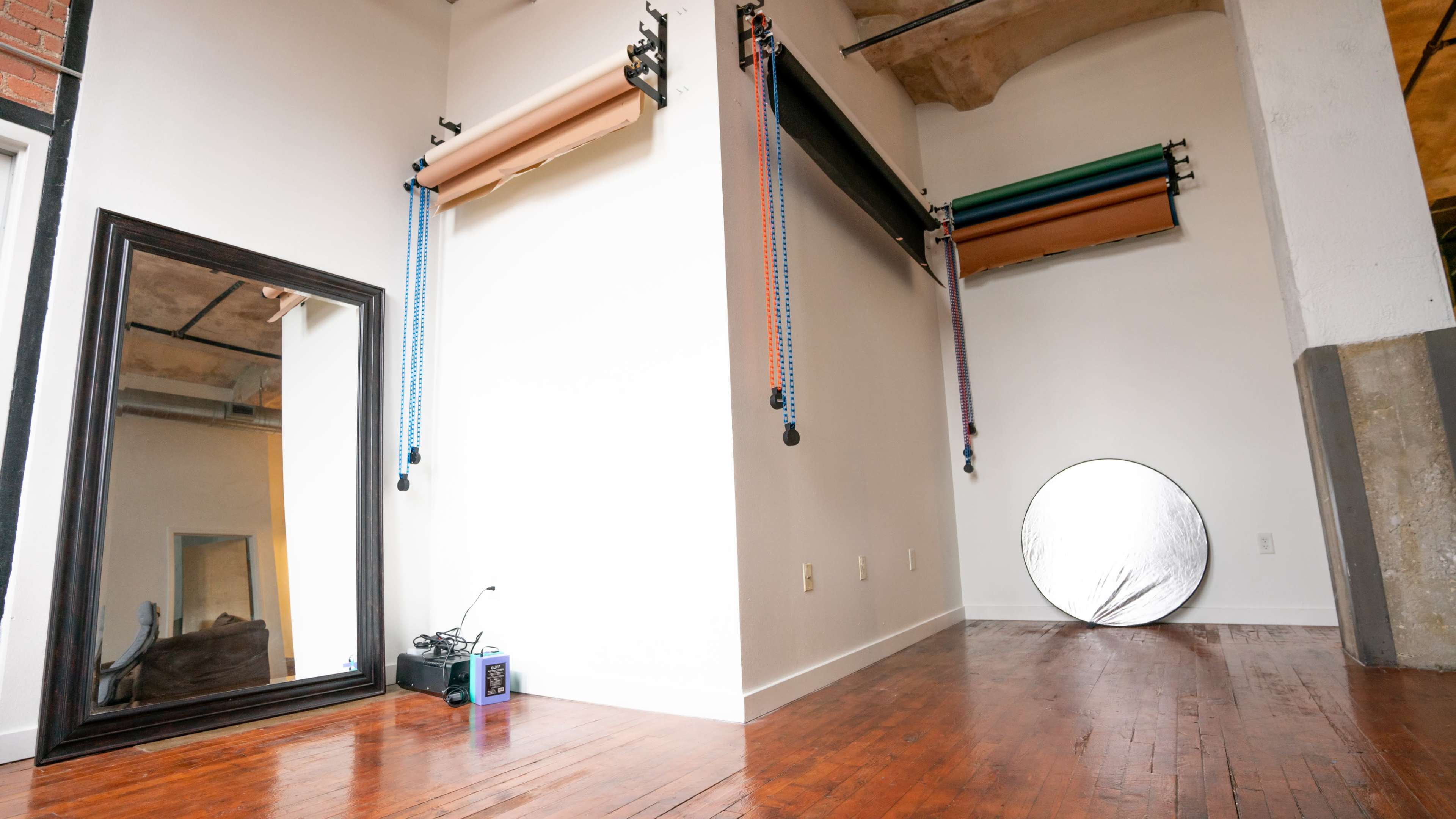A studio space with a large mirror on one wall, backdrops mounted and hanging on the ceiling, and a circular reflector on the hardwood floor.