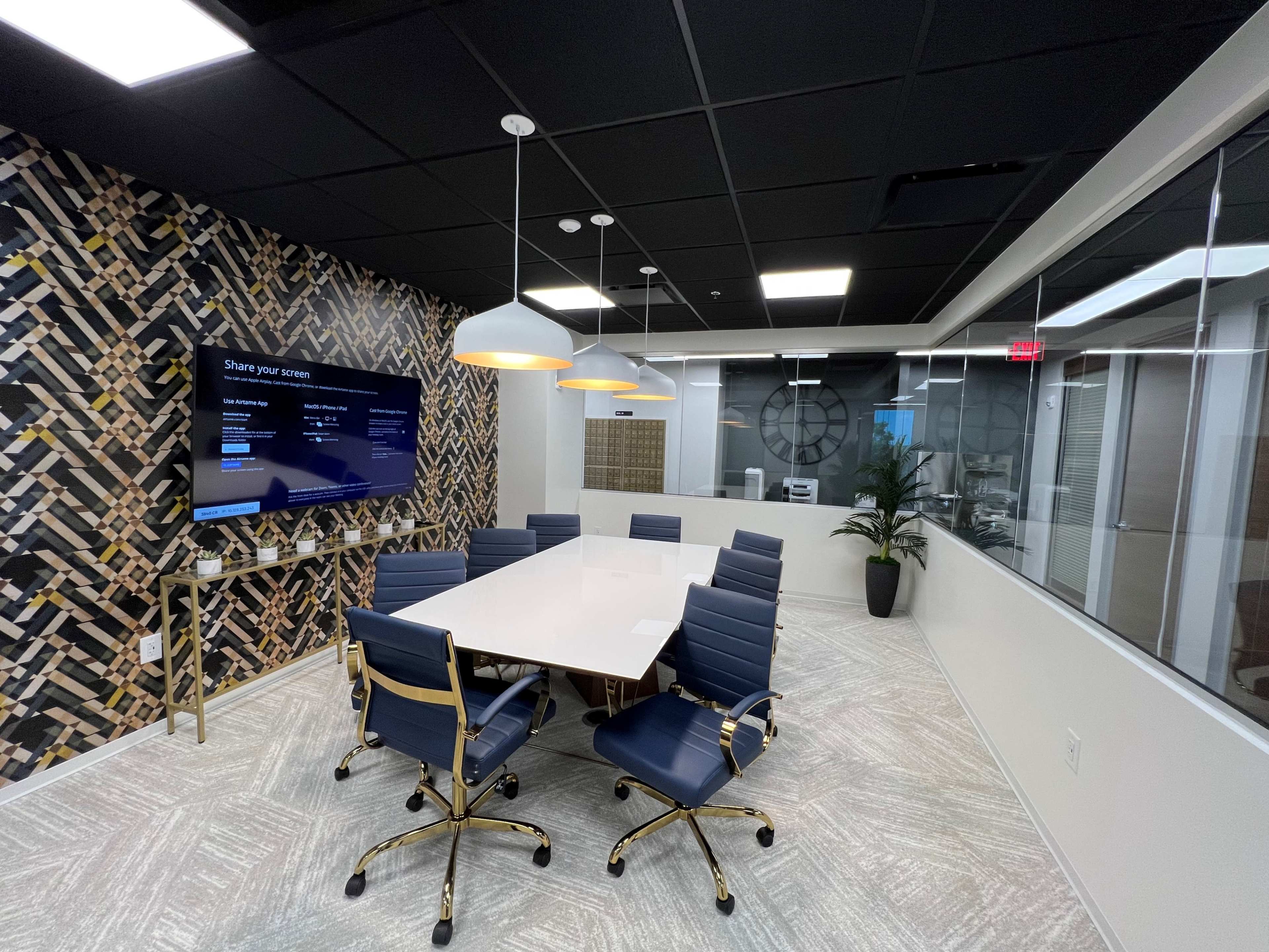 The image shows a modern conference room with a large white table surrounded by blue chairs, a large wall-mounted screen, and a patterned accent wall.