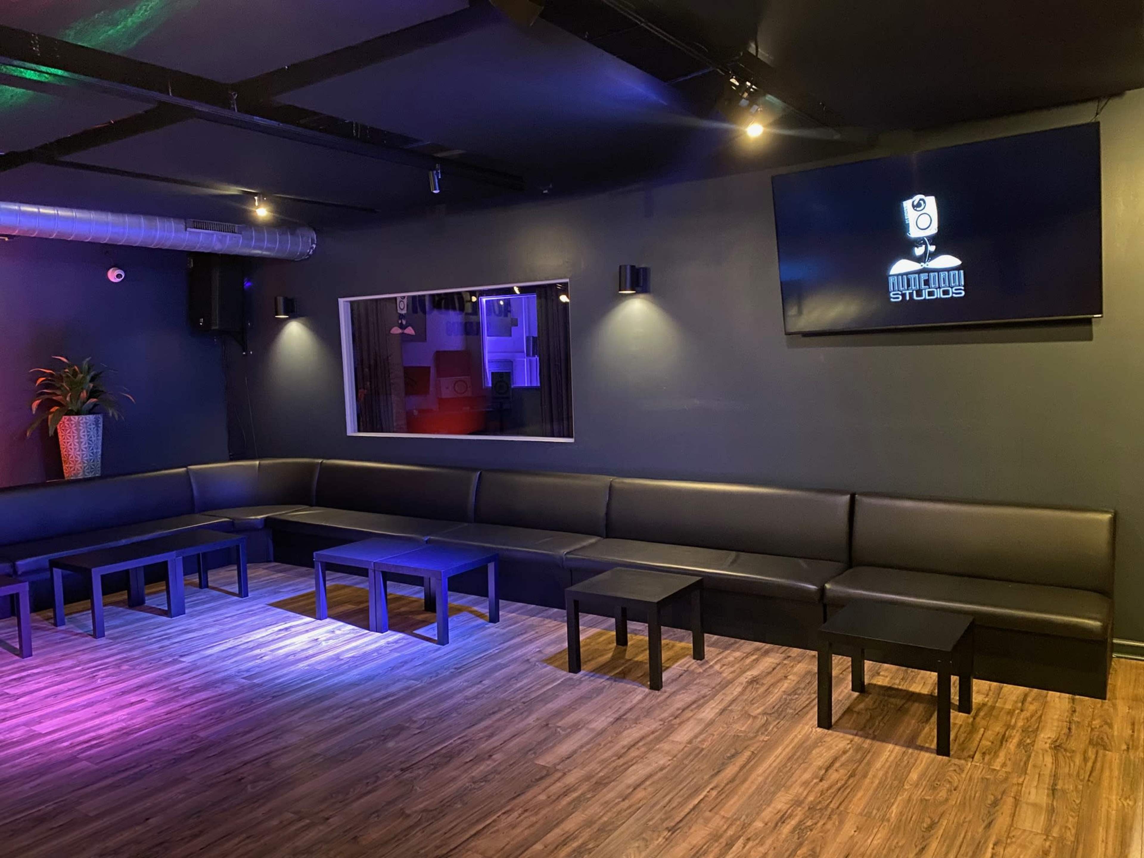 A dimly lit lounge area with a black leather bench, several small tables, and a large screen displaying a logo on the wall.