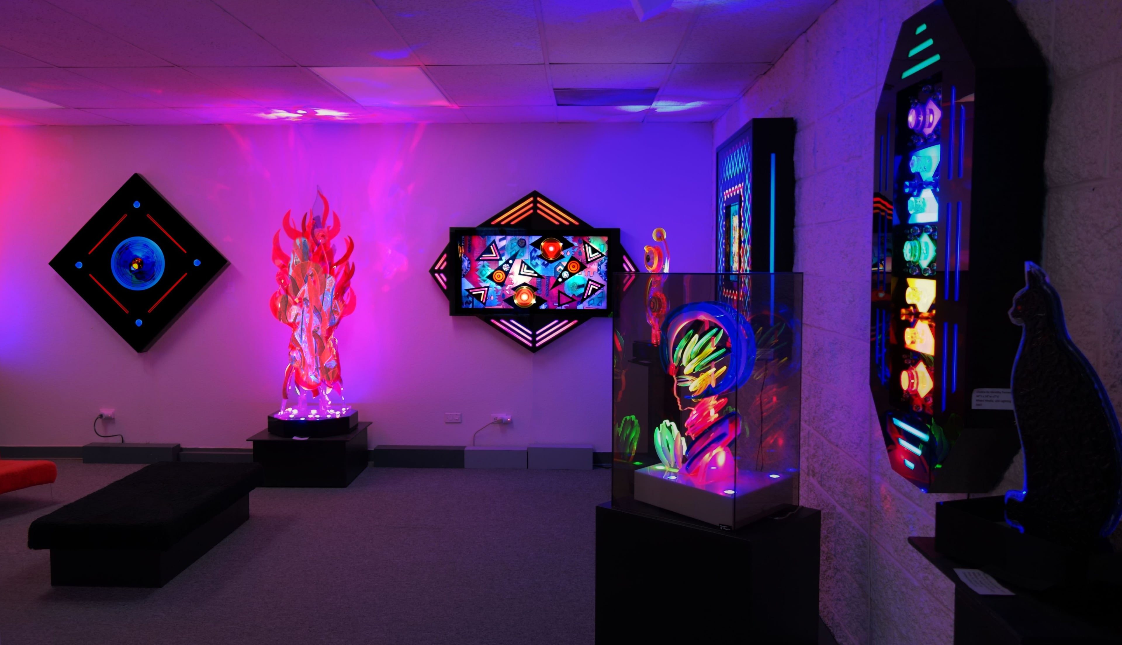 An art gallery featuring brightly colored, illuminated installations and digital displays on the walls, accompanied by a glass display case containing vibrant sculptures.