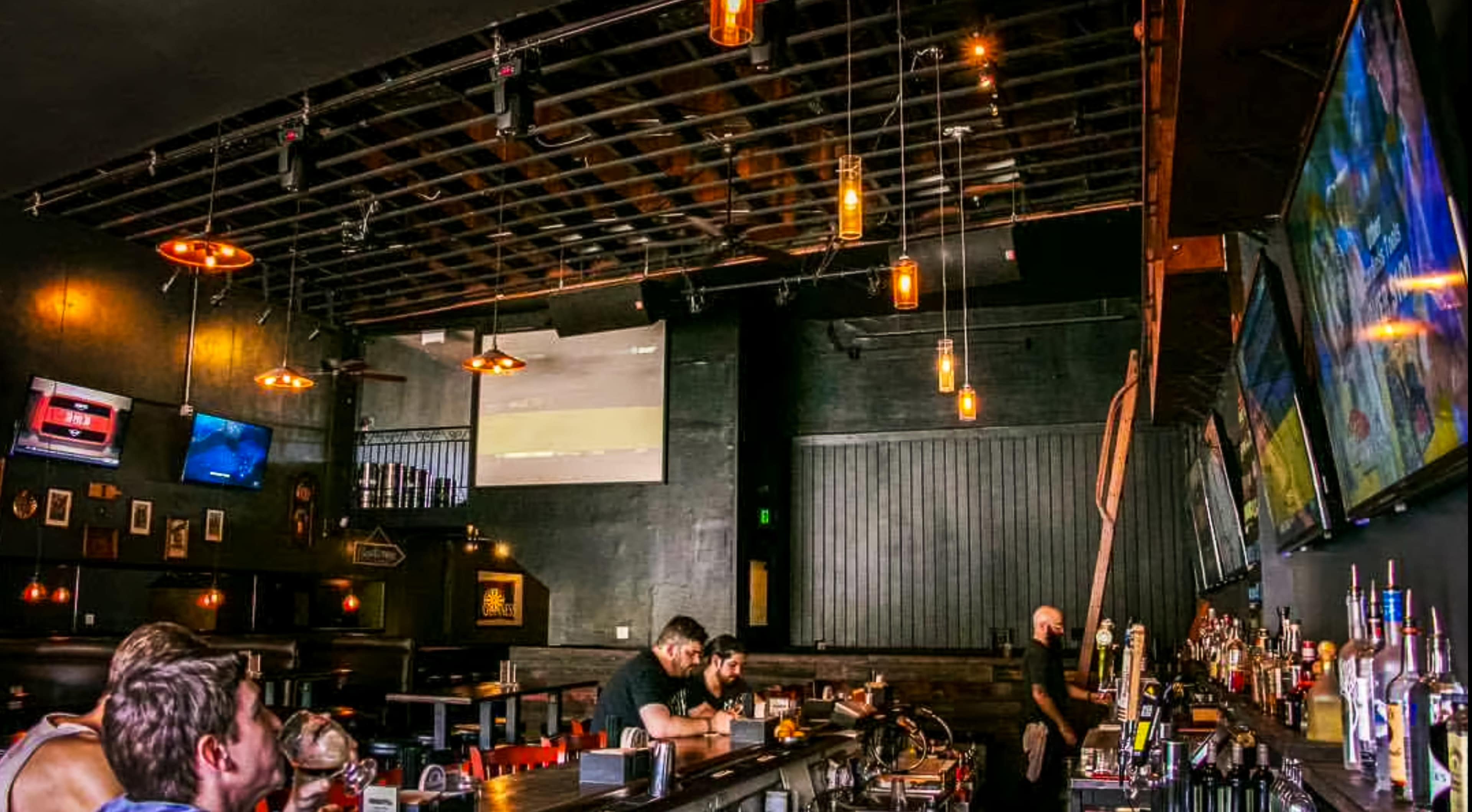 The image shows a dimly lit bar with patrons sitting at the counter, multiple screens displaying sports events, and illuminated pendant lights hanging from the ceiling.