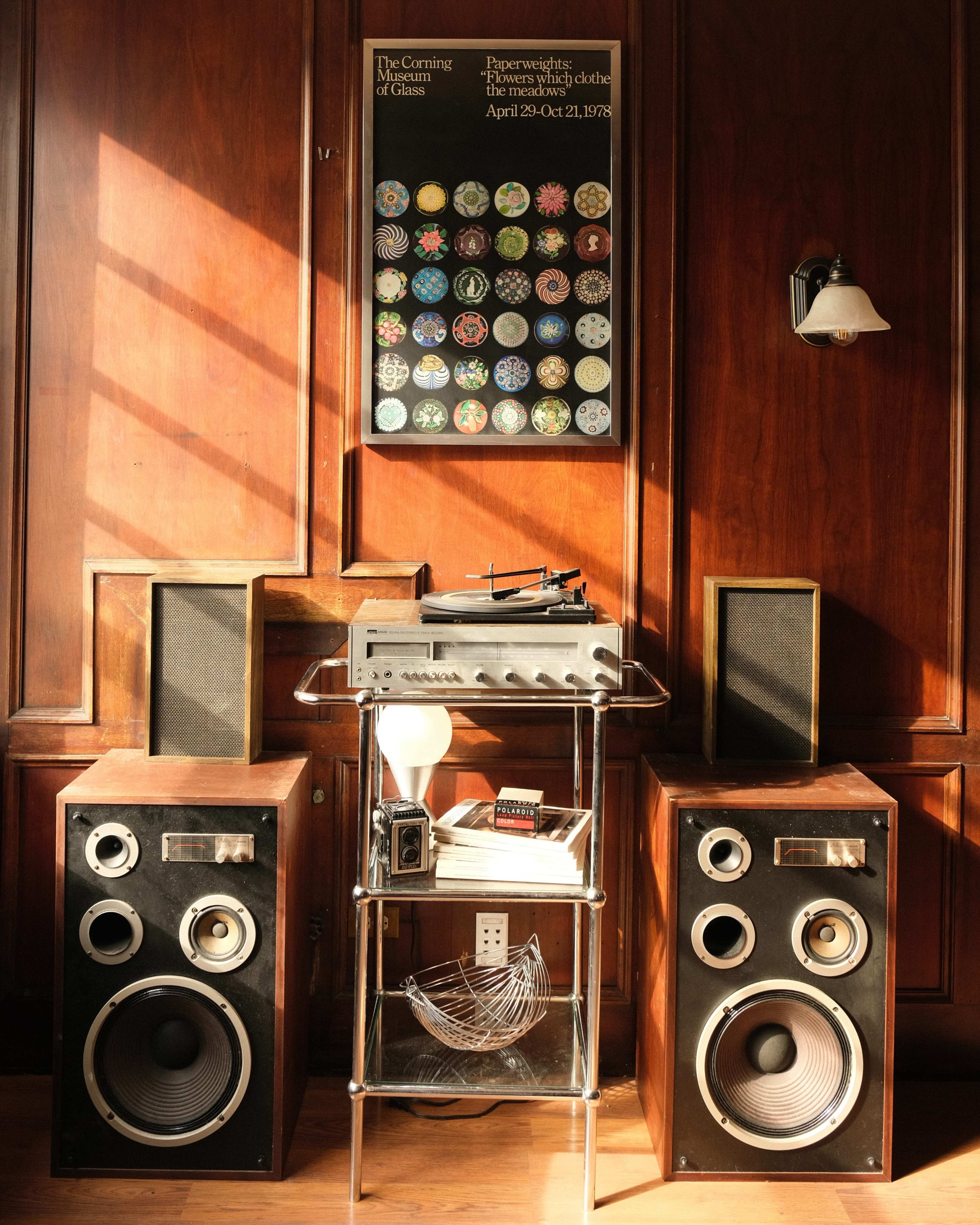 The image shows a vintage audio setup with two large speakers, a record player on a metal stand, and a colorful poster hanging on a wooden wall.