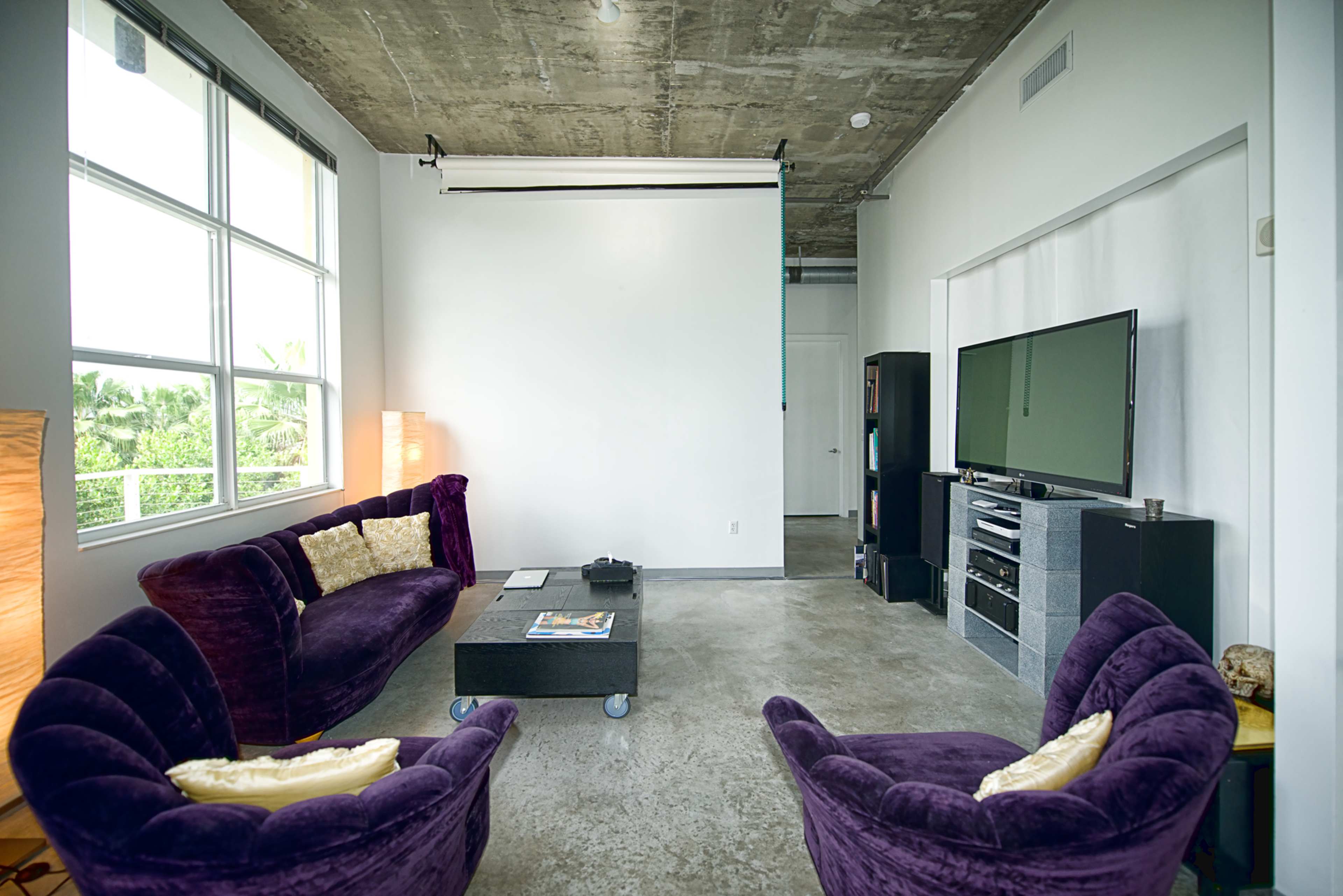 The image shows a modern living room with purple velvet furniture, including a sofa and two armchairs, against a backdrop of concrete walls and large windows.