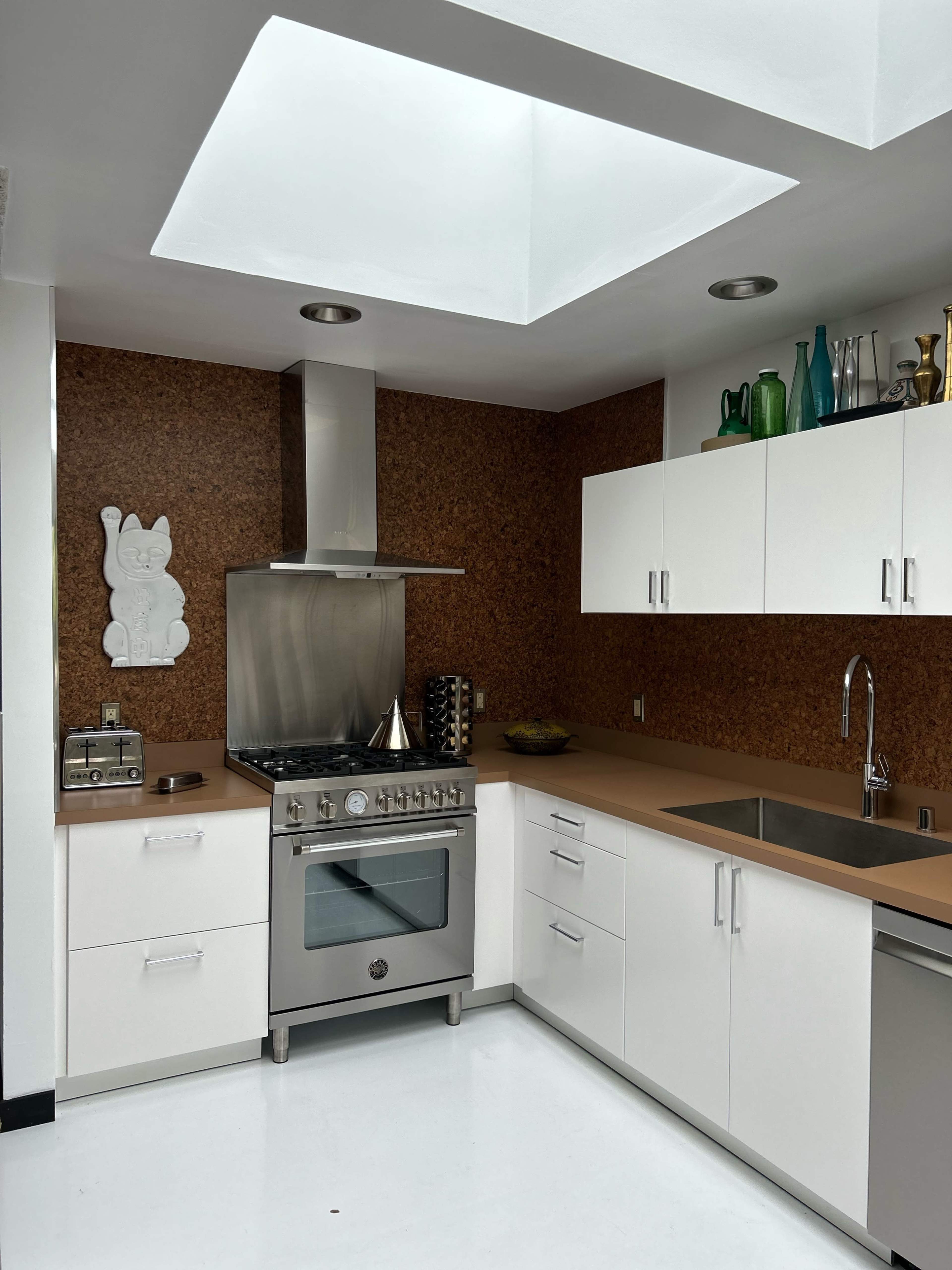 The kitchen features modern stainless steel appliances, white cabinets, and a cork accent wall, illuminated by a skylight above.