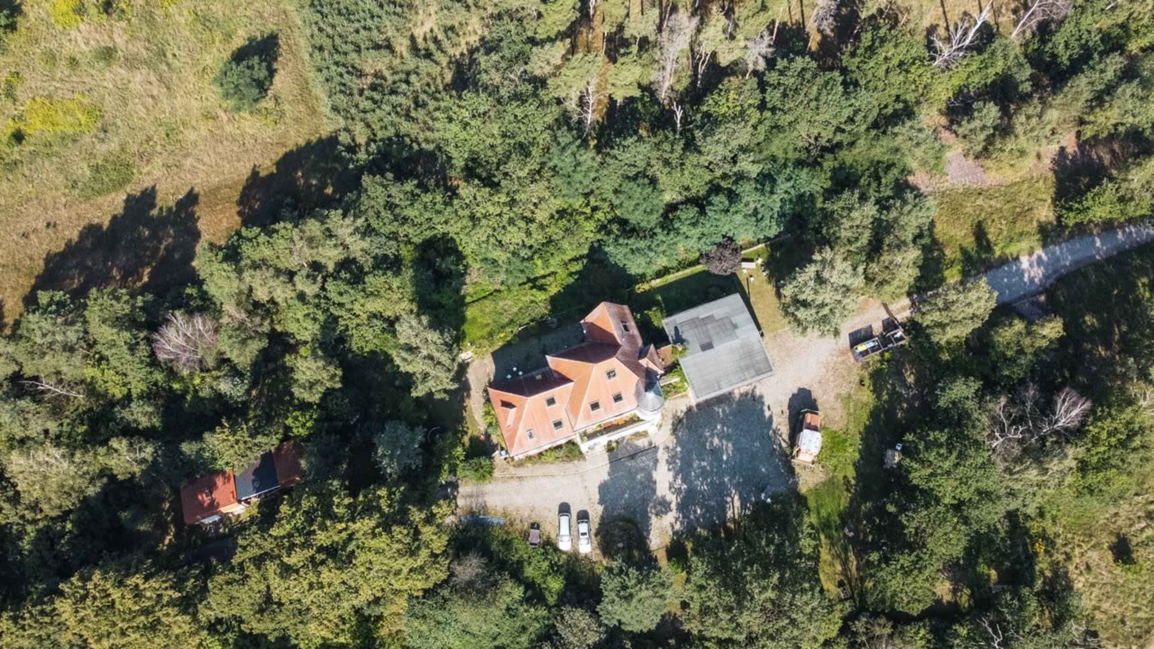An aerial view shows a house surrounded by dense greenery, with a gravel driveway leading to the property and additional parked vehicles nearby.