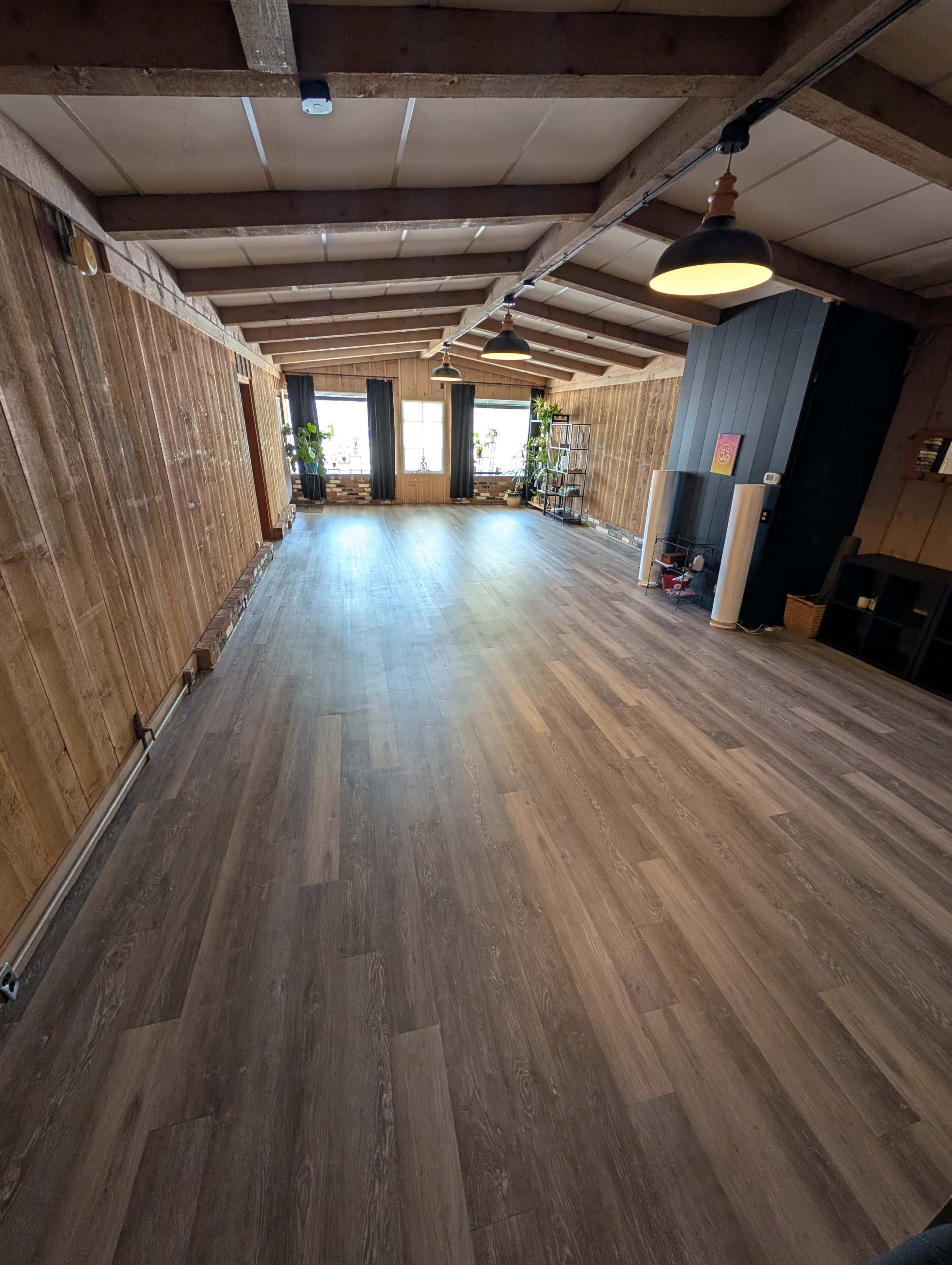 The image shows a spacious, empty room with wooden walls and a laminate floor, illuminated by natural light from large windows.