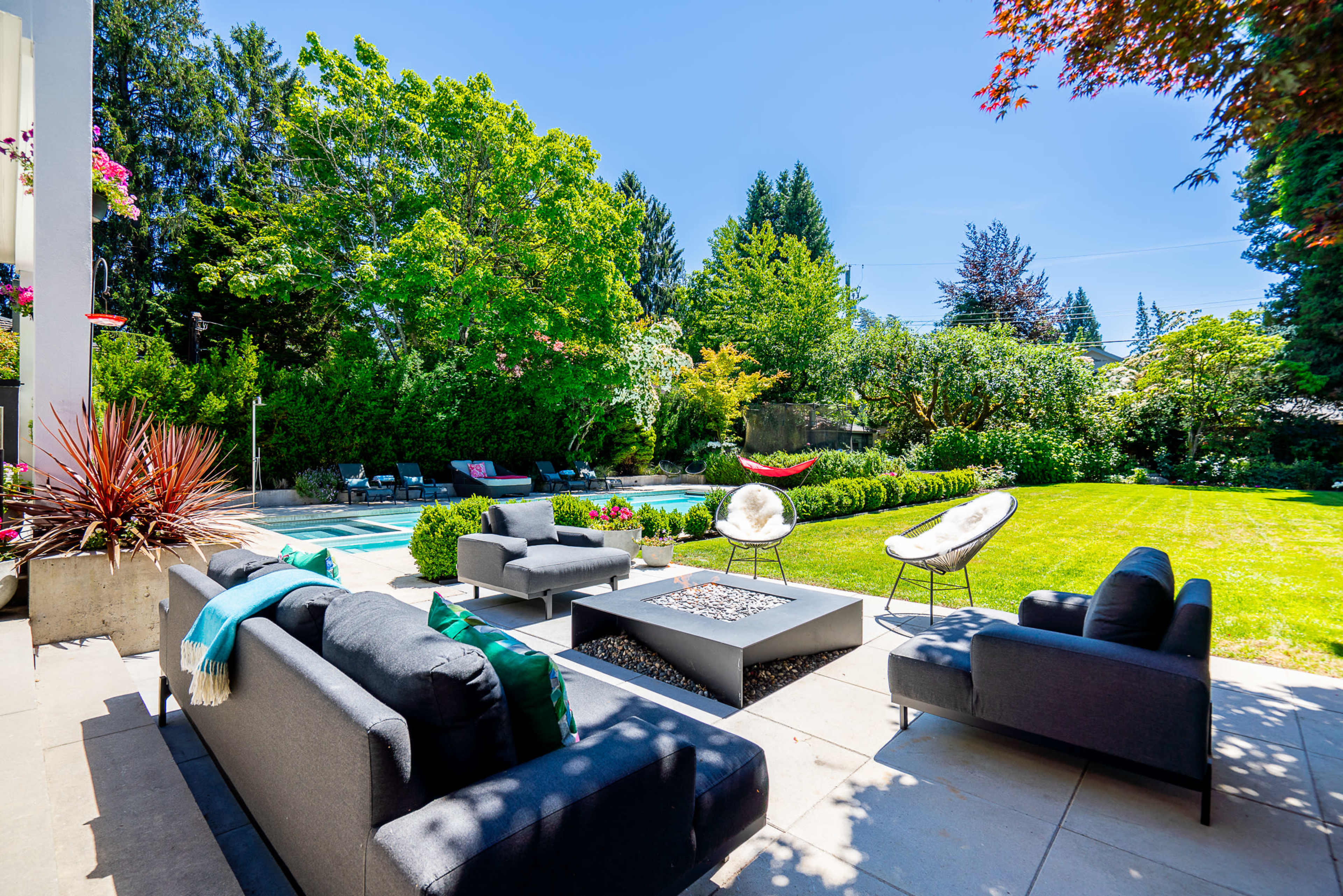 The image shows a landscaped backyard with a swimming pool, seating area featuring sofas and a fire pit, and well-maintained greenery.