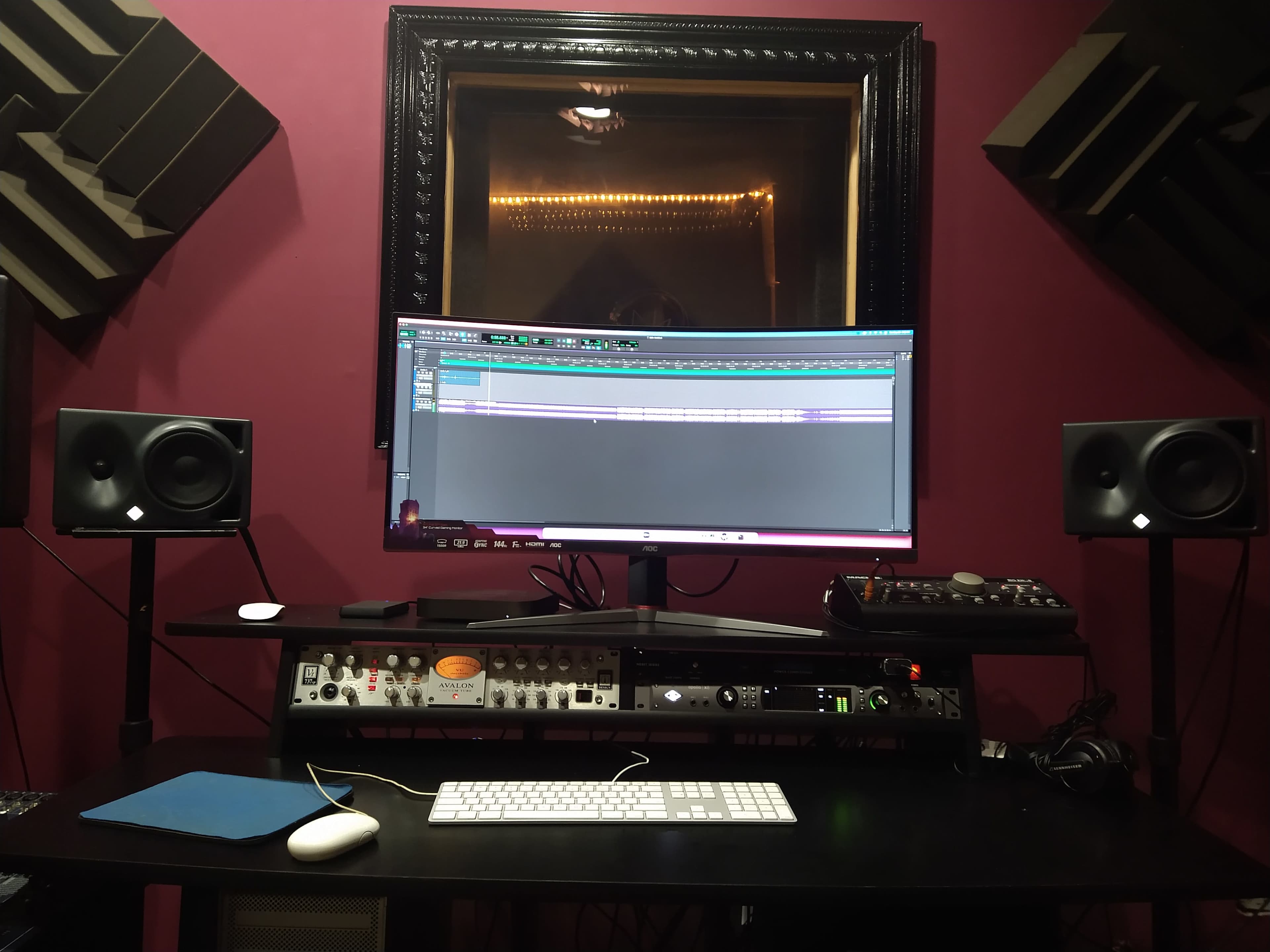 A music production desk features a large monitor displaying editing software, flanked by studio speakers and an audio interface on a dark tabletop.
