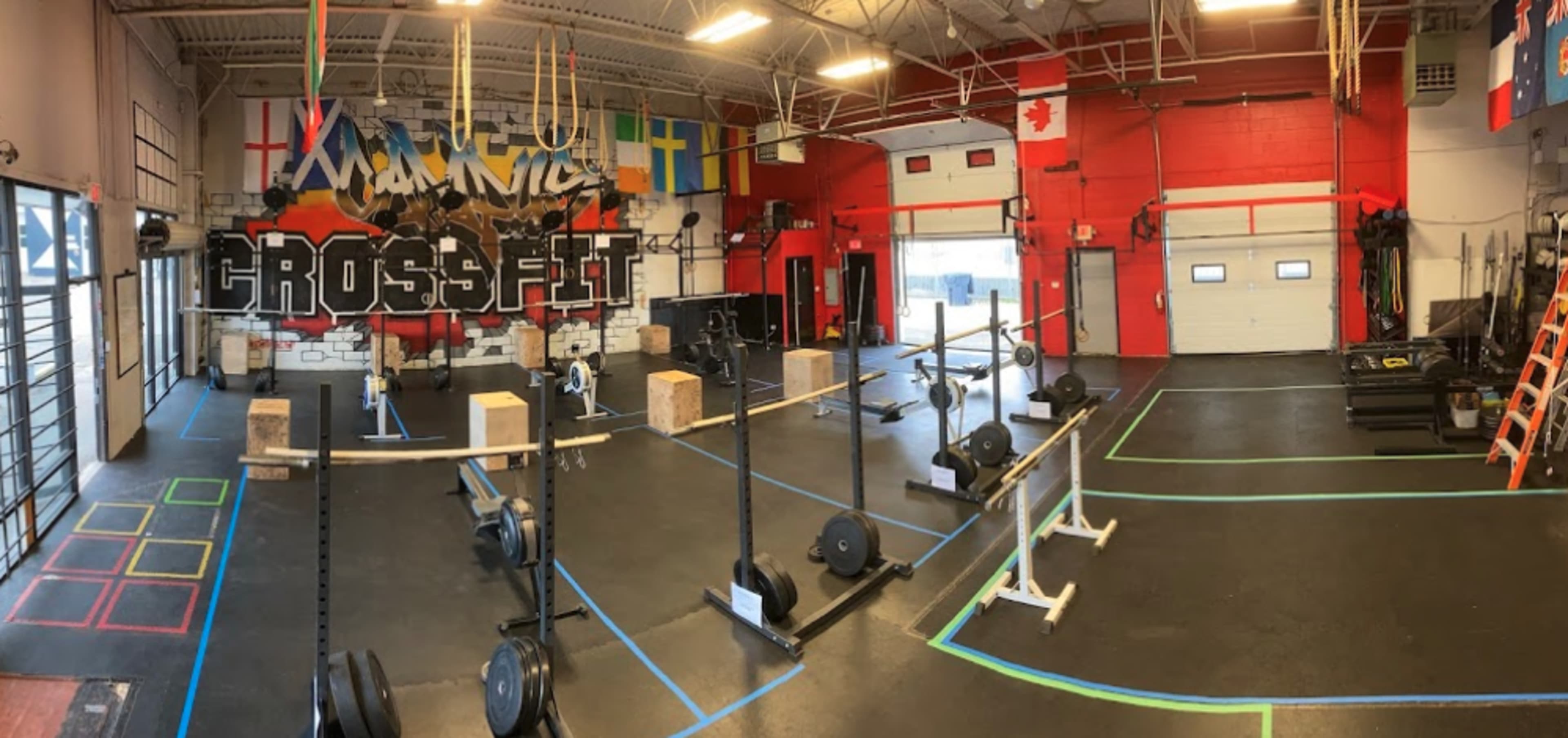A CrossFit gym with various workout equipment arranged on a black rubber floor, illuminated by overhead lights and decorated with flags and artwork on the walls.