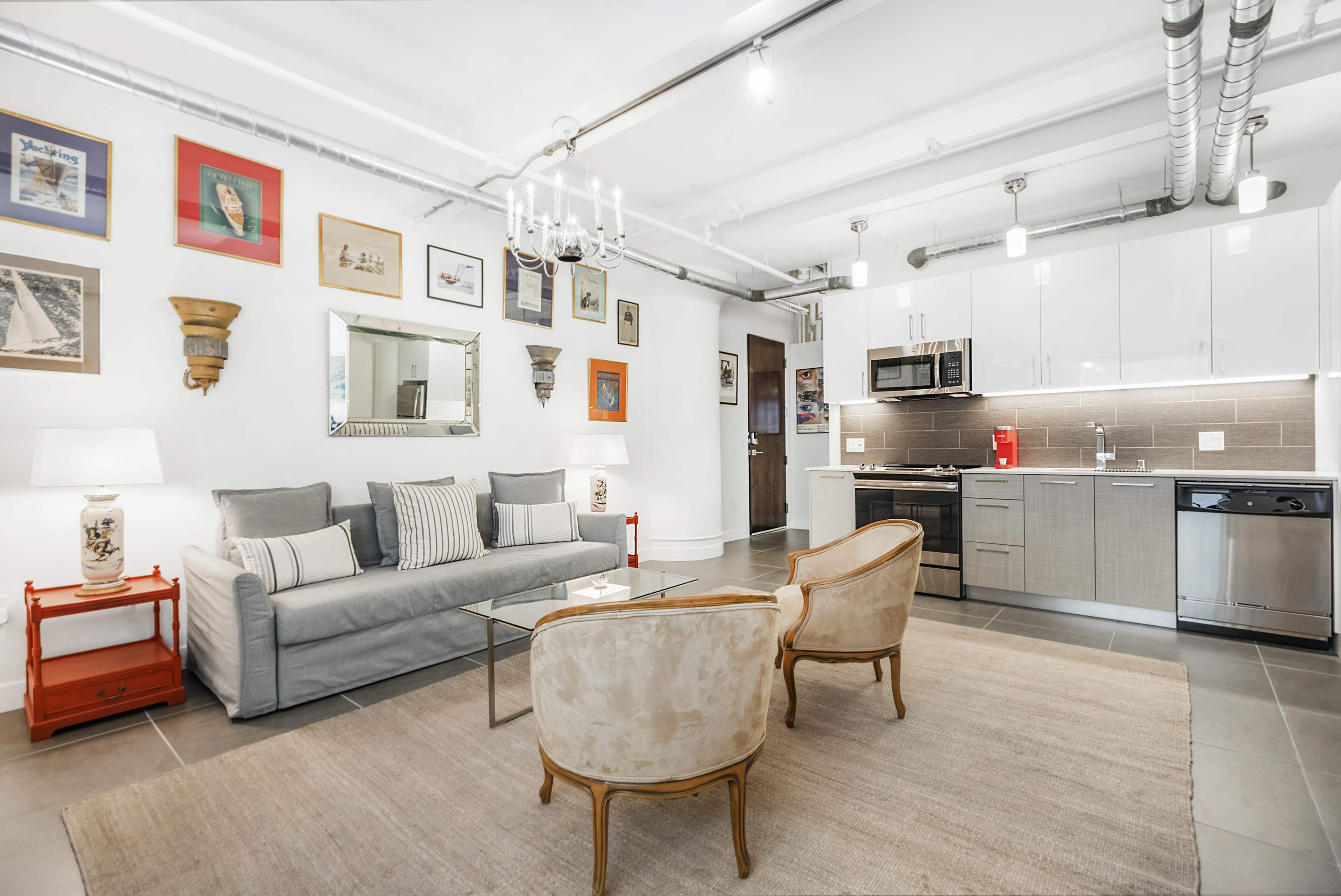 The image shows a modern living area with a gray sofa, two upholstered chairs, a coffee table, and a kitchenette with stainless steel appliances.