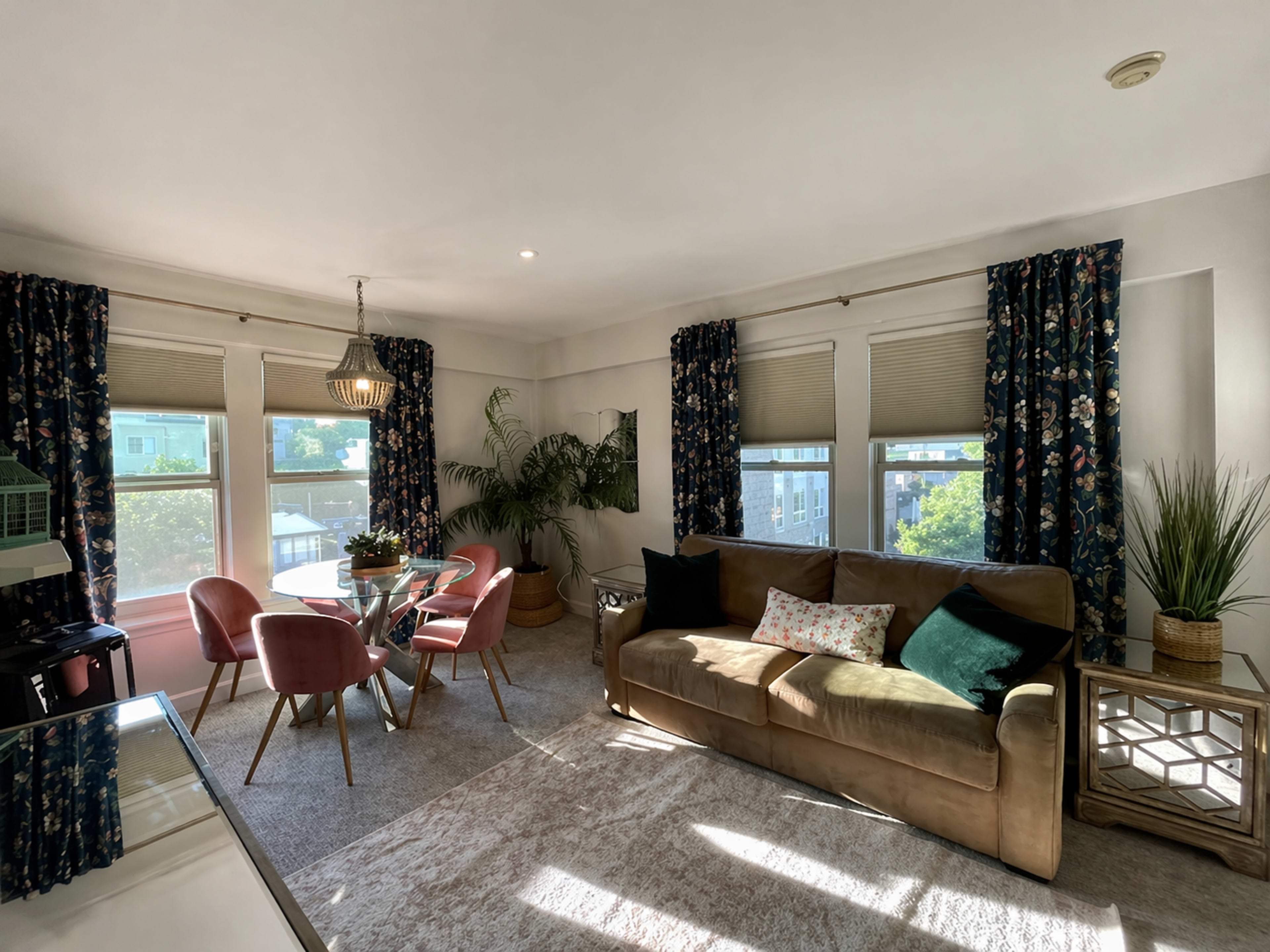 A cozy living room features a brown sofa, a glass dining table with pink chairs, and large windows covered with floral curtains.
