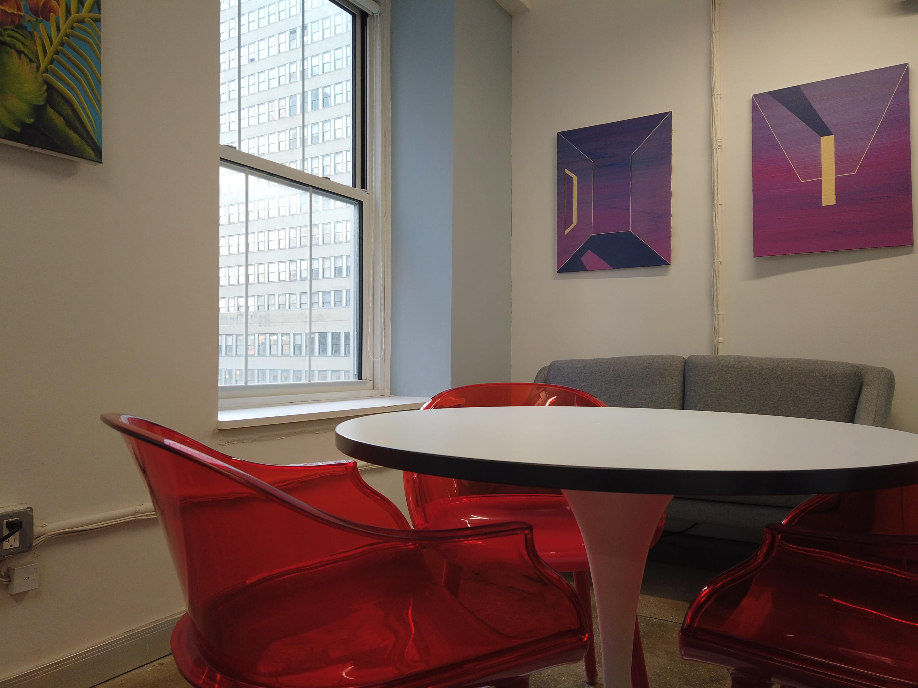 The image shows a small meeting space with a circular table surrounded by three transparent red chairs and a gray sofa, alongside two abstract wall paintings and a view of a city building through the window.
