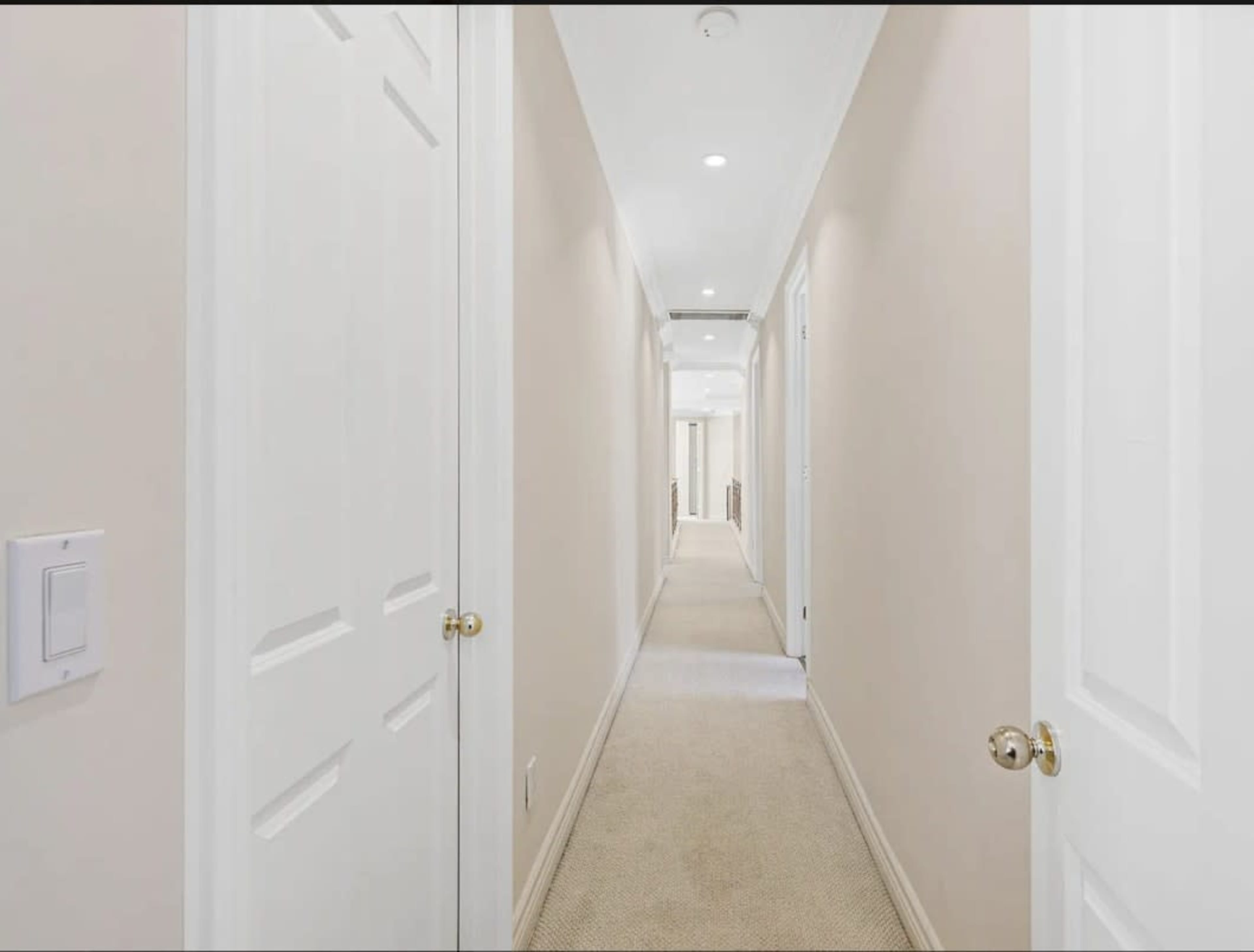 A clean, beige hallway with doors on each side and carpeted flooring extends into the distance, illuminated by overhead lights.