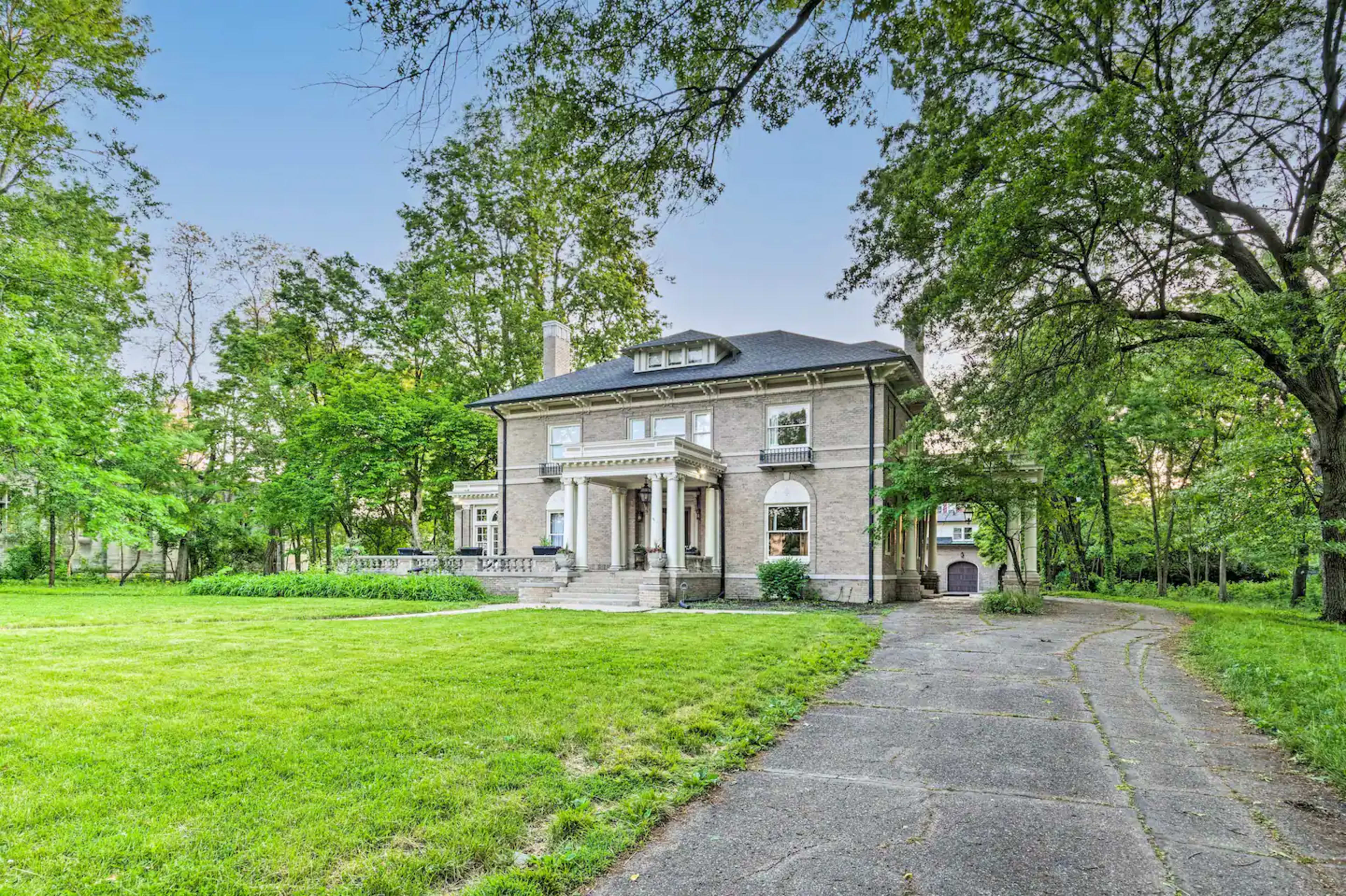 The image shows a large, two-story house with a stone facade, surrounded by green trees and a grassy yard, with a driveway leading up to it.