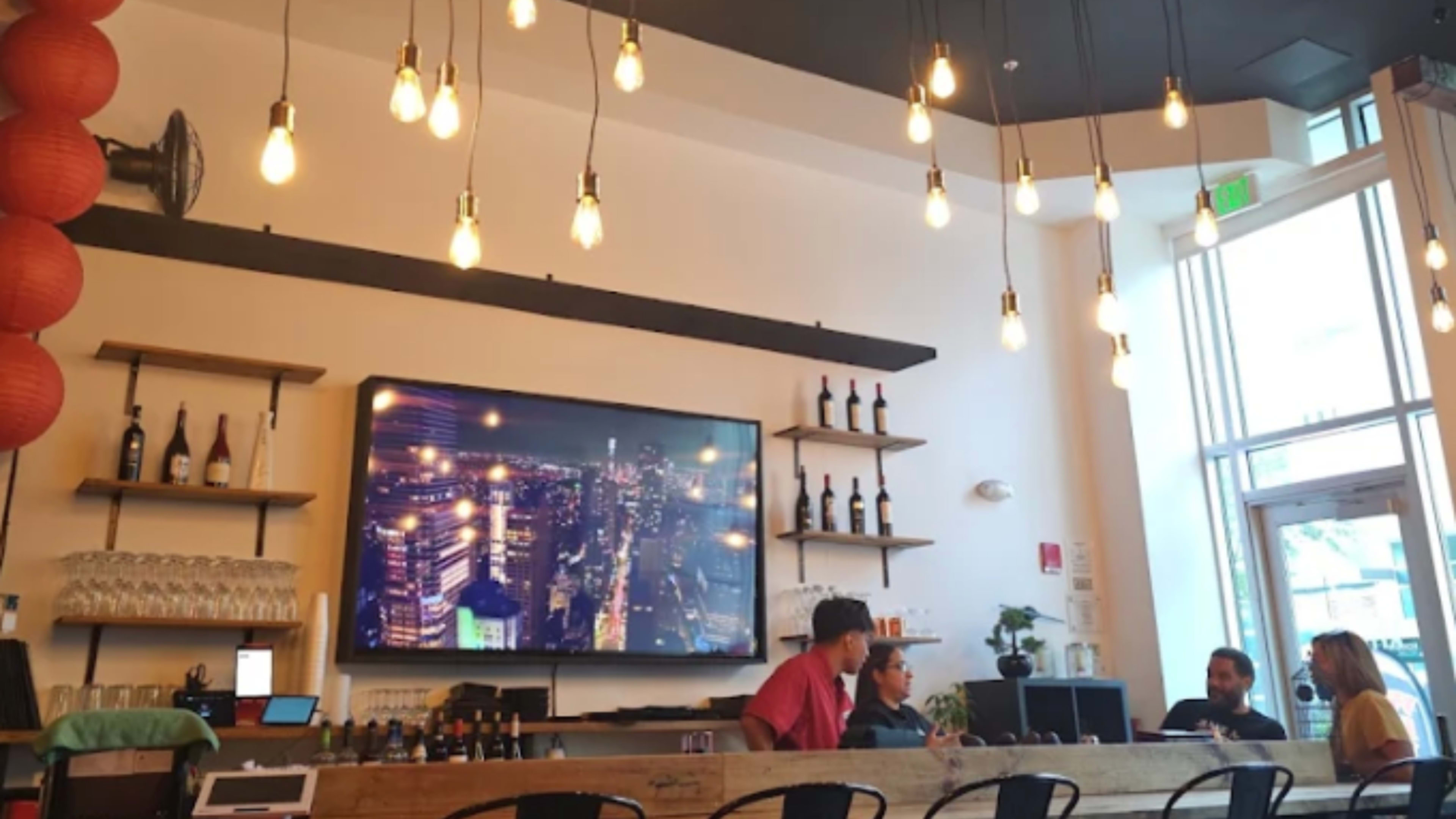 The image shows a modern bar area with hanging light fixtures, a large screen displaying a city skyline, and patrons seated at the counter.