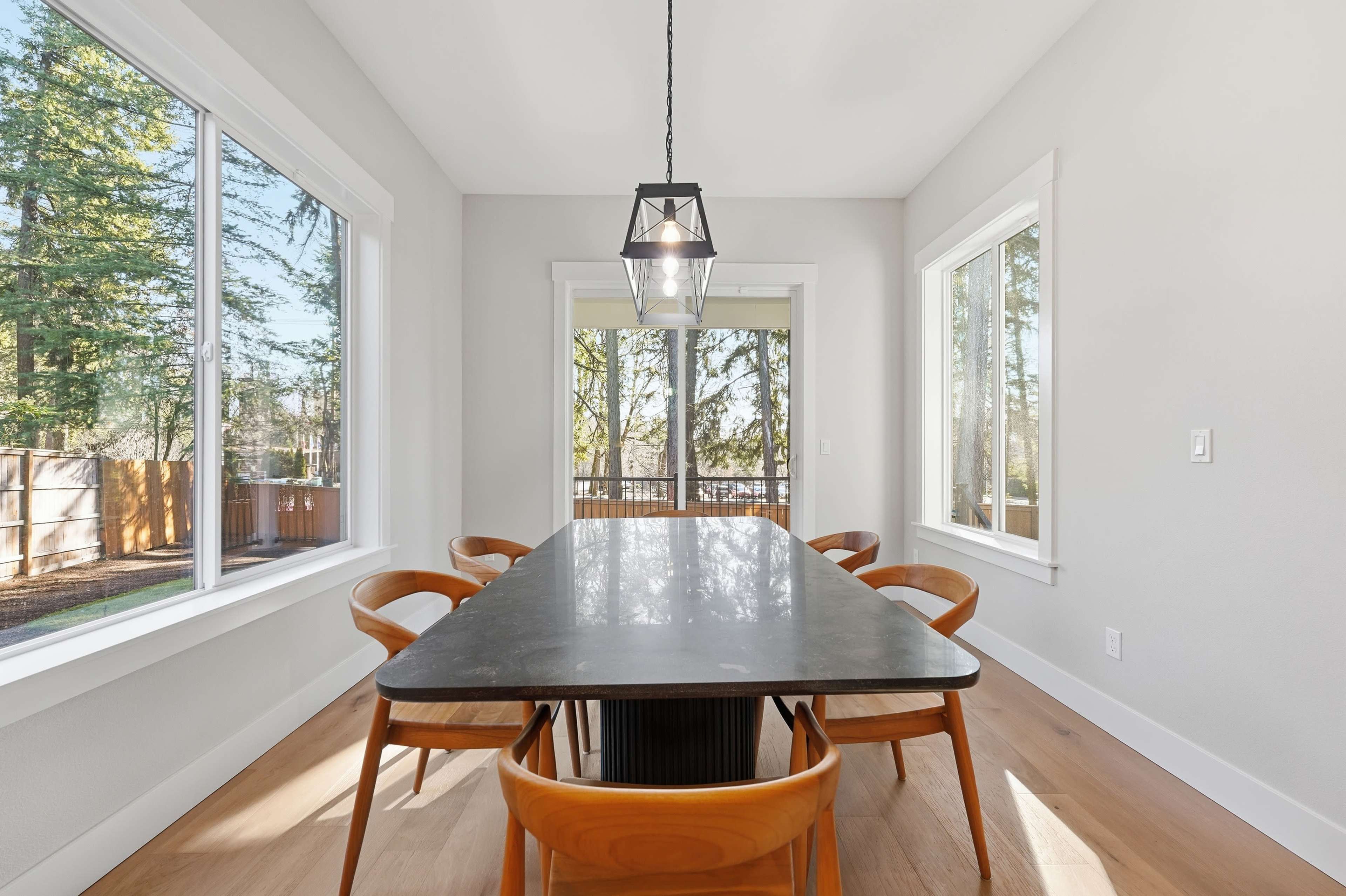 A dining area features a long rectangular table surrounded by wooden chairs, with large windows letting in natural light and a view of trees outside.