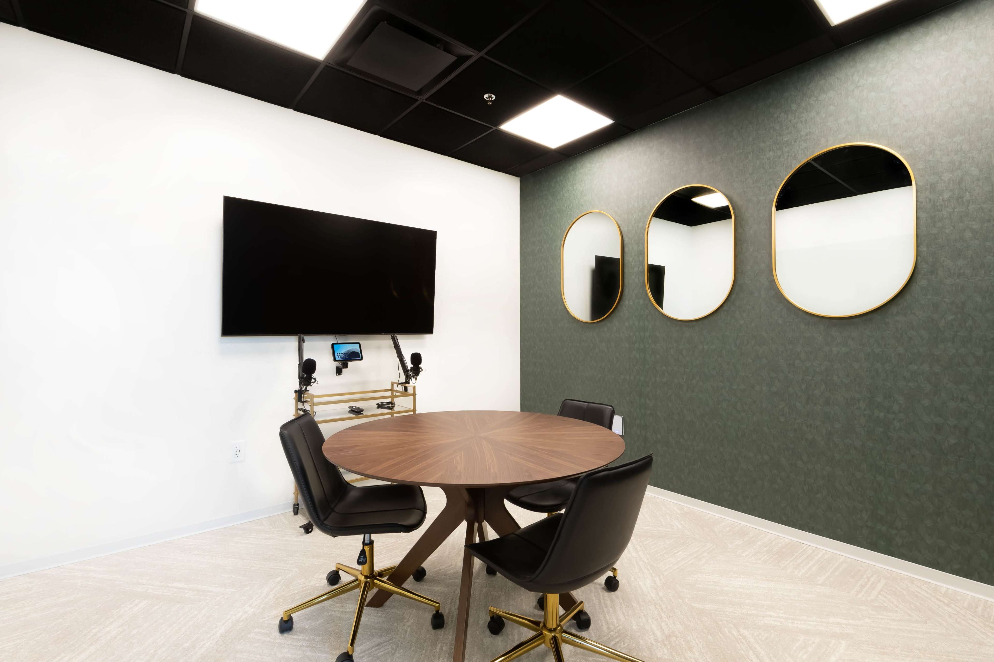 A conference room features a round wooden table with black chairs, a large TV mounted on the wall, and three circular mirrors.