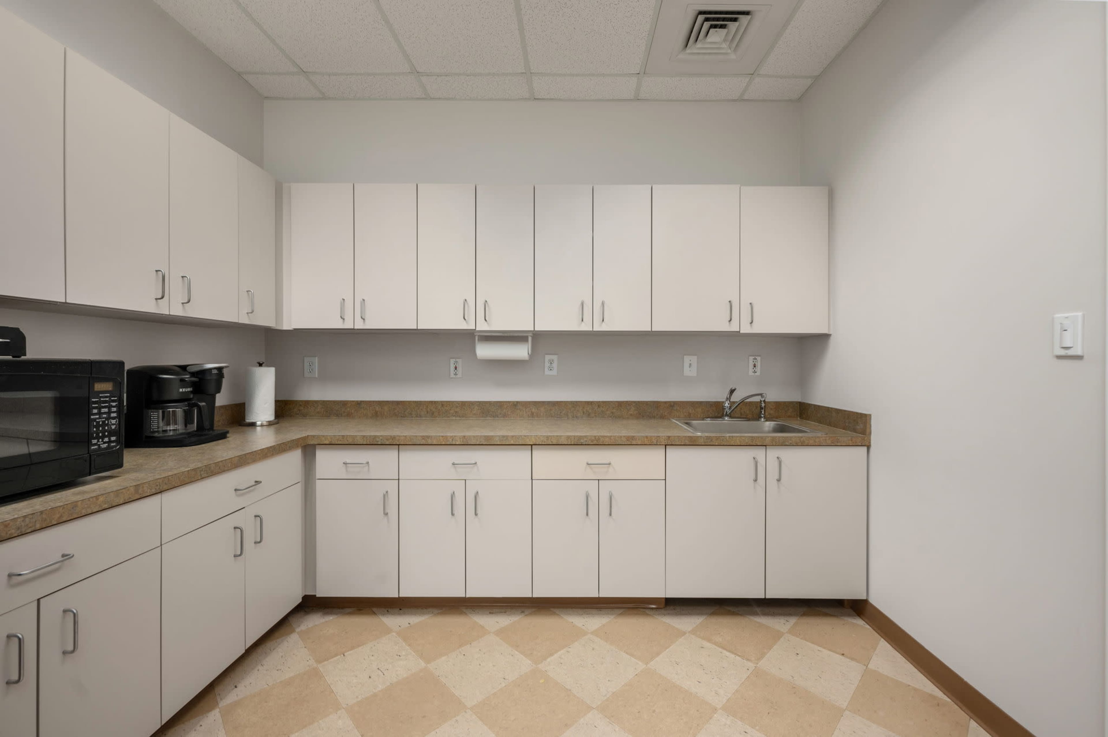 A clean, organized kitchen with light-colored cabinets and a countertop, featuring a microwave and a sink.