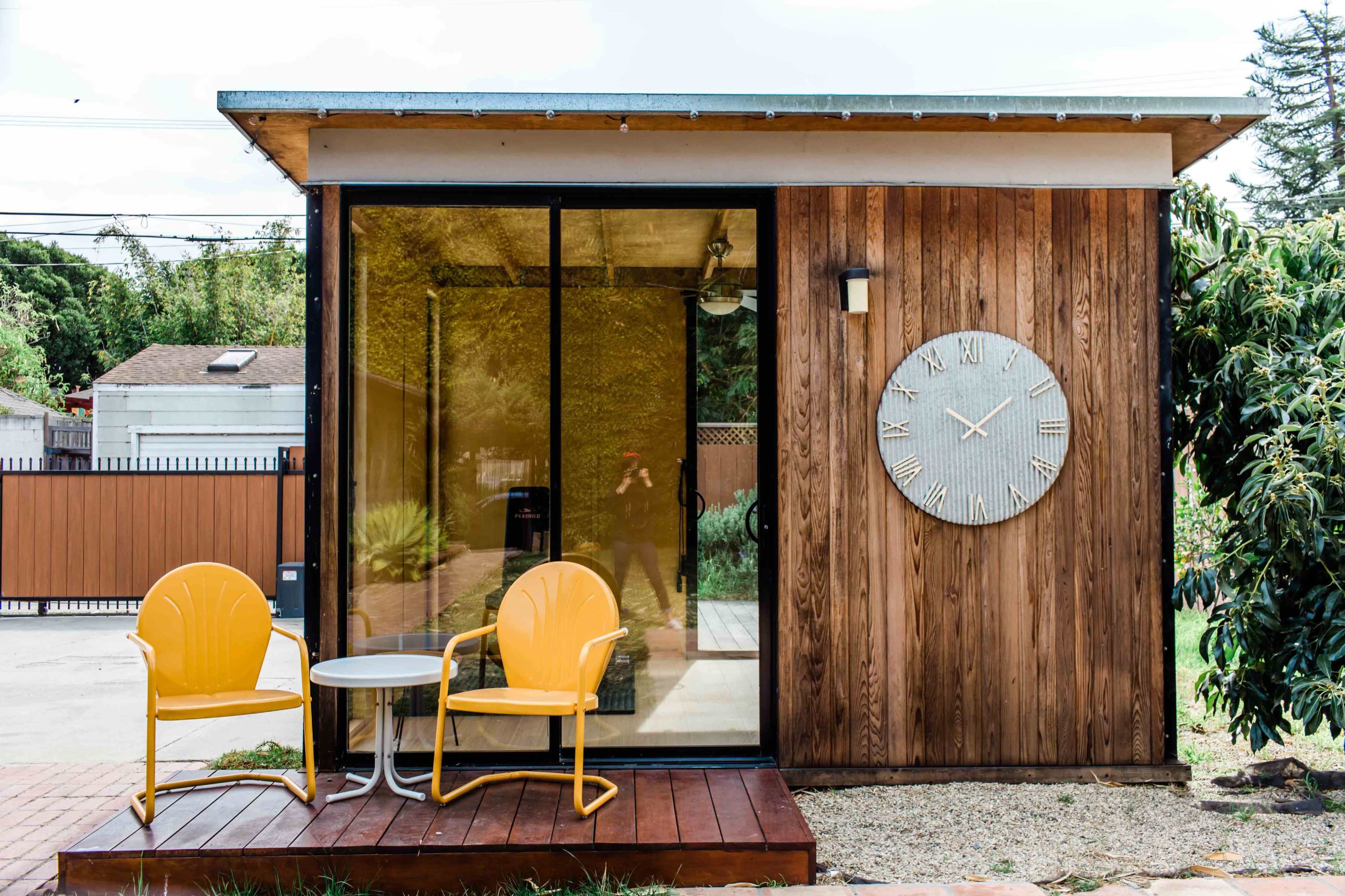 The image shows a small wooden structure with a large clock on the wall, featuring two yellow chairs and a round table on a wooden deck in front.