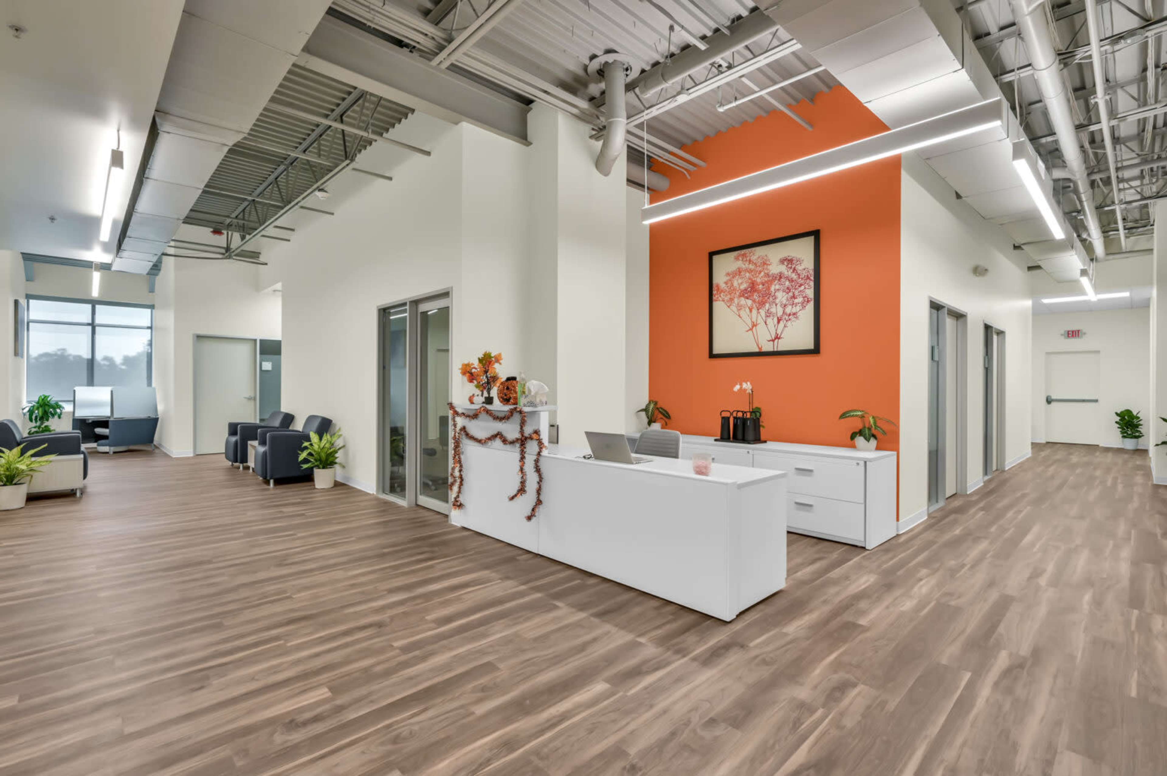 The image shows a modern office reception area with a white front desk, an orange accent wall, and wooden flooring, complemented by plants and seating arrangements.