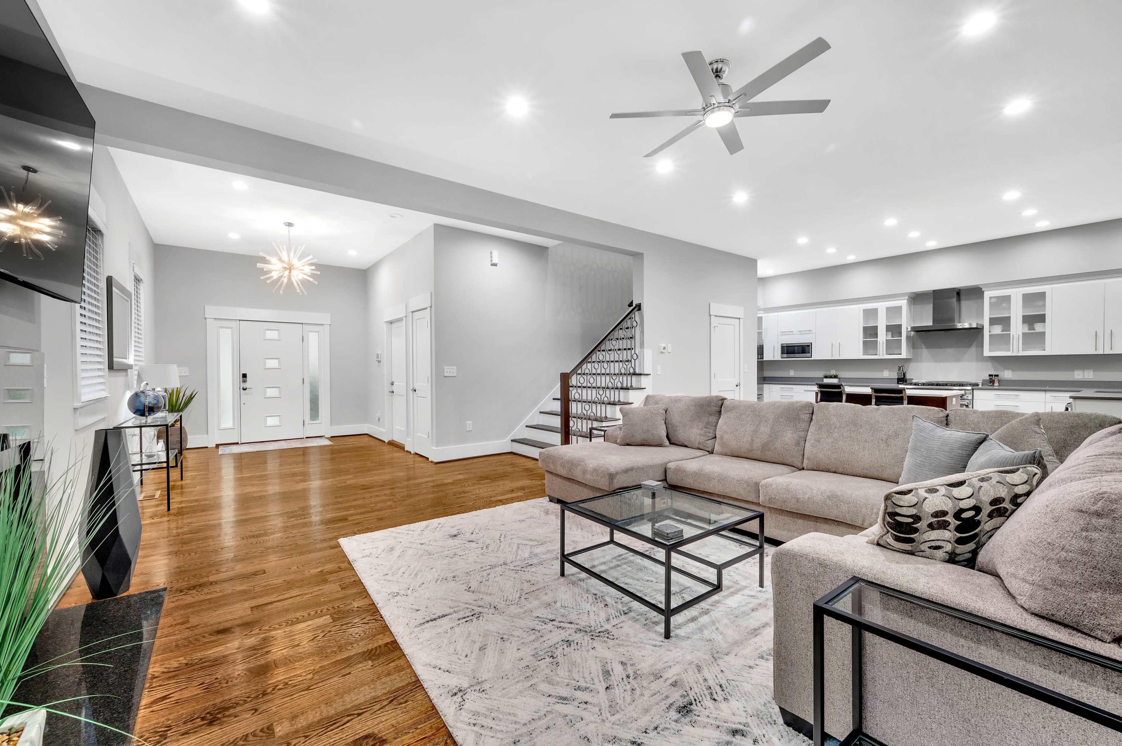 A modern living room features a spacious layout with a sectional sofa, a glass coffee table, and a view into an open kitchen area.