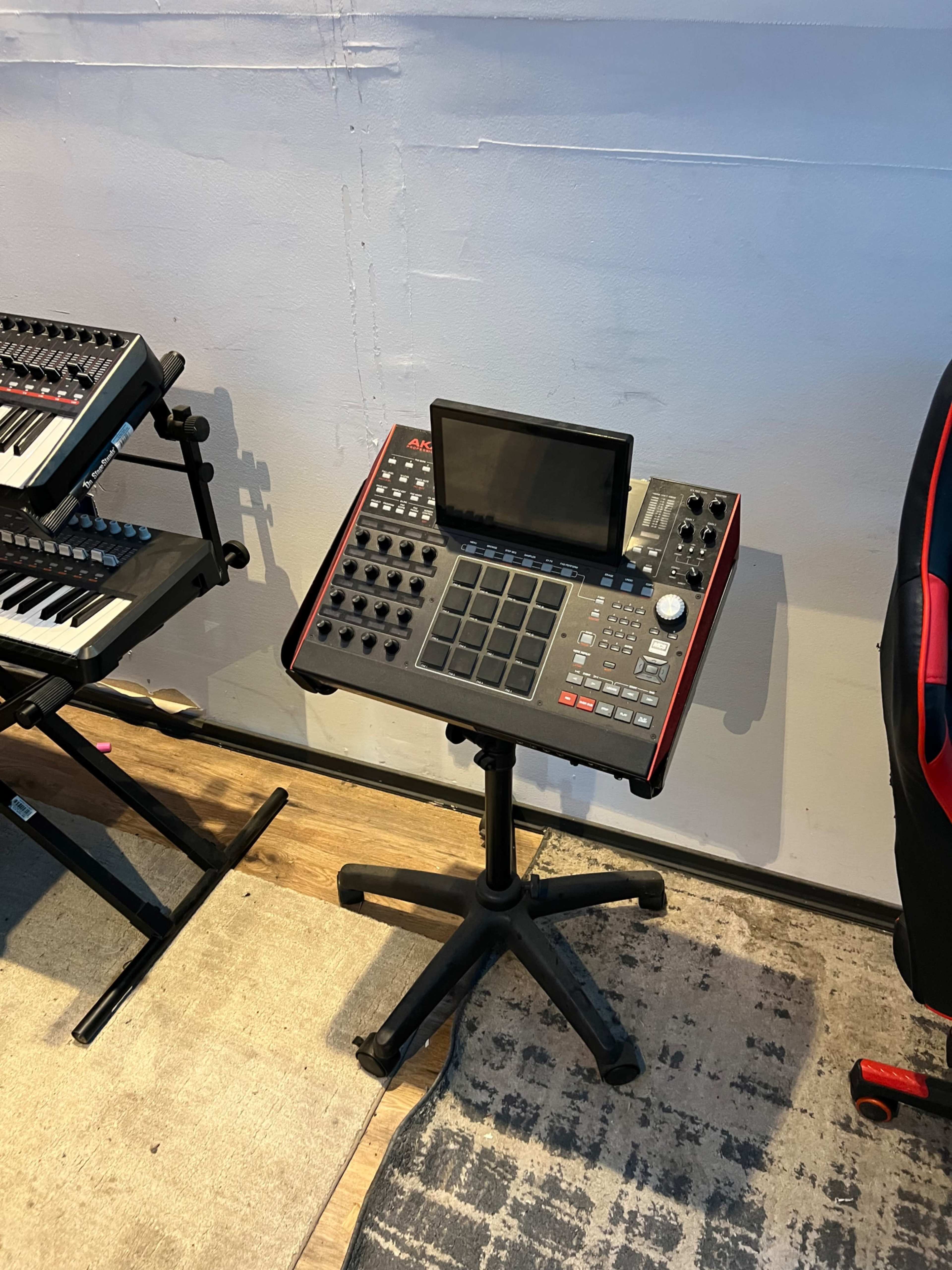 A music production workstation featuring an Akai MPC performance pad mounted on a black adjustable stand, positioned next to two keyboards.