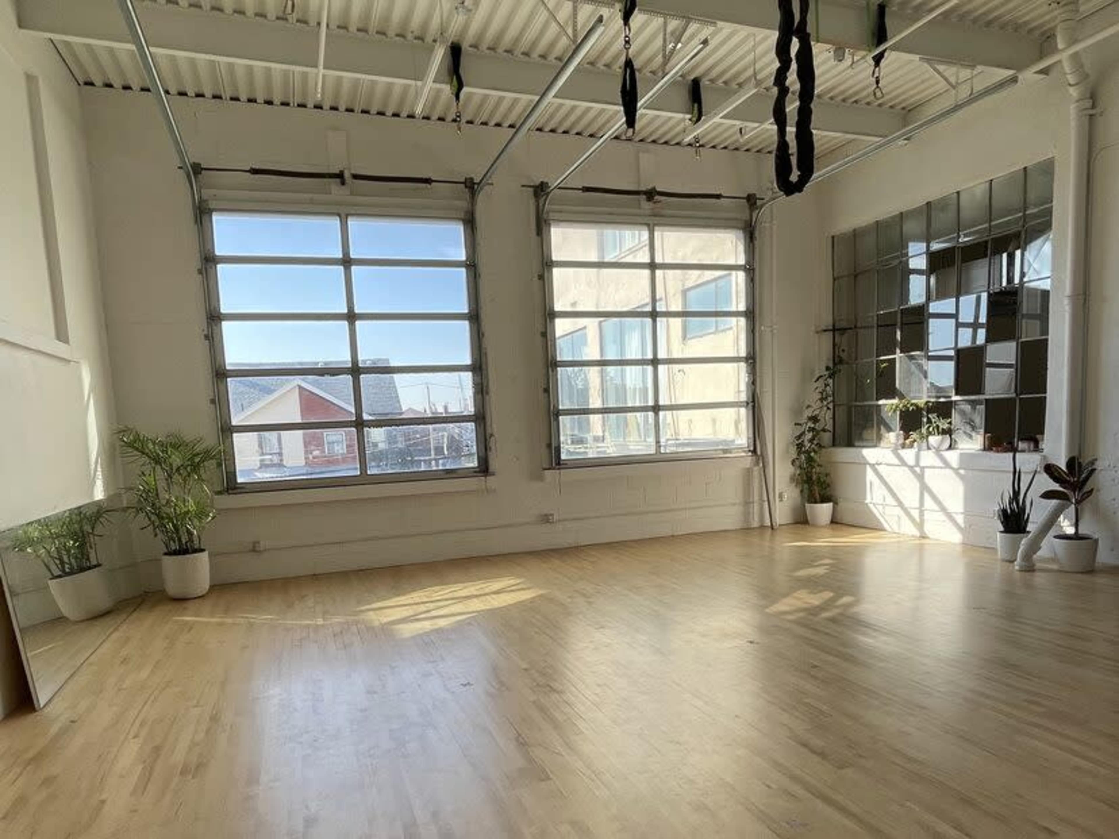 A spacious, well-lit room with large windows, wooden flooring, and potted plants along the walls.
