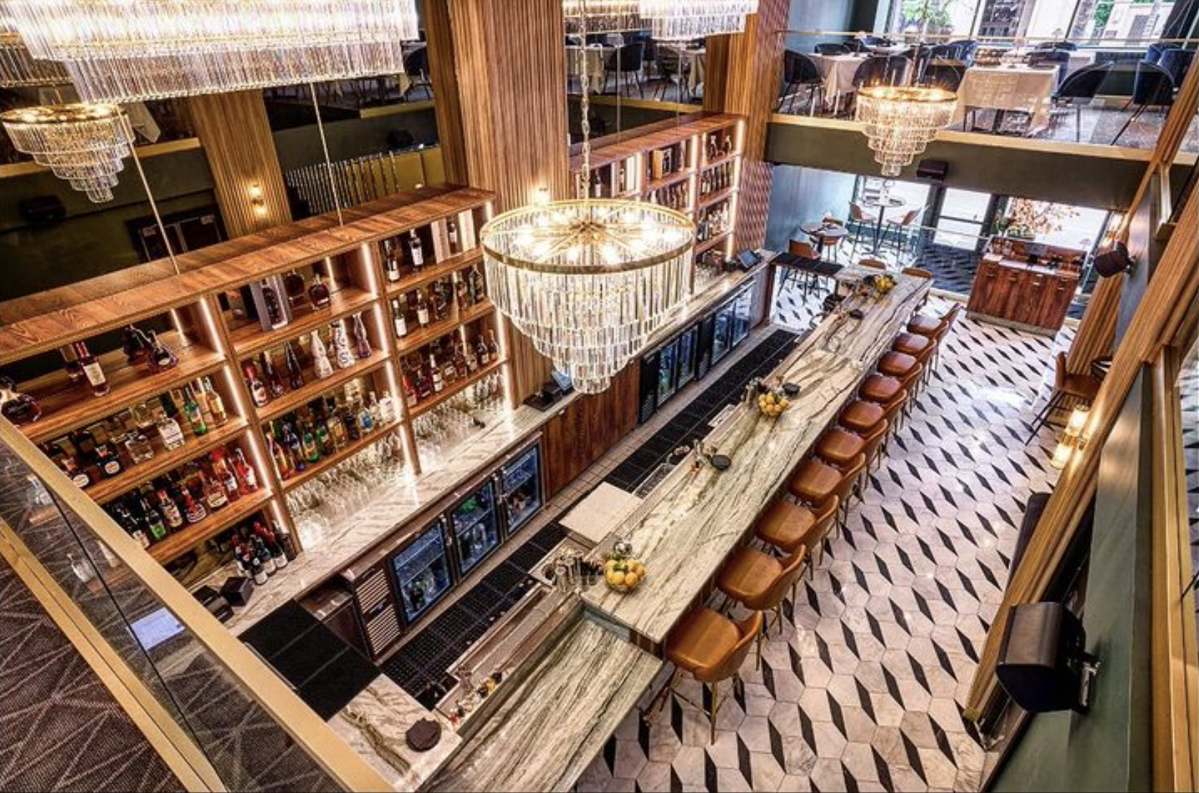 A spacious bar with a long marble countertop, surrounded by seating and illuminated by large chandeliers.