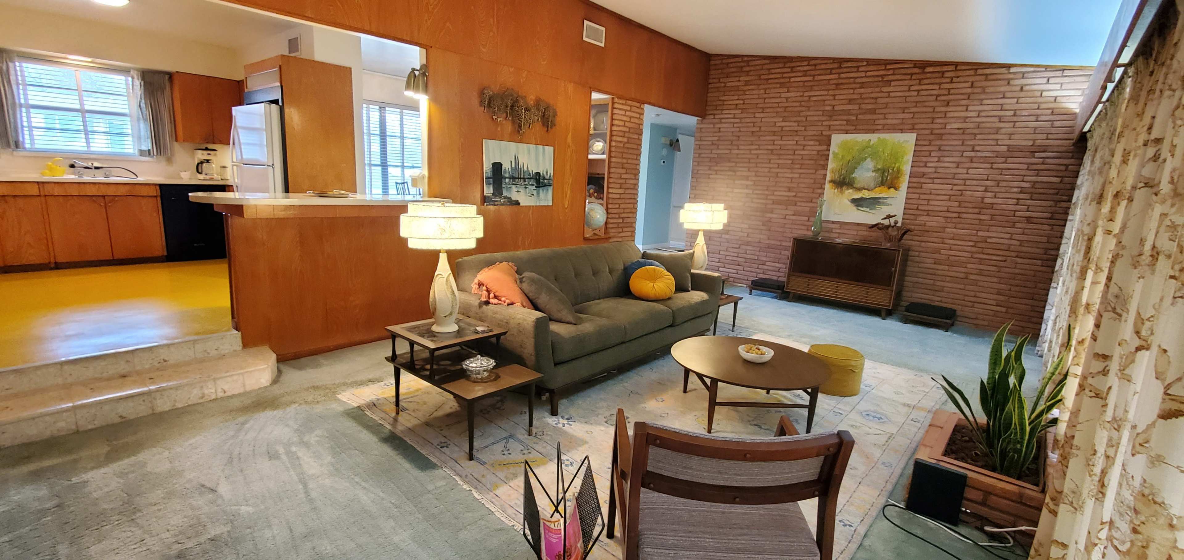 A modern living room with a green sofa, two table lamps, a coffee table, and a view into a kitchen area featuring wooden cabinets and a yellow floor.
