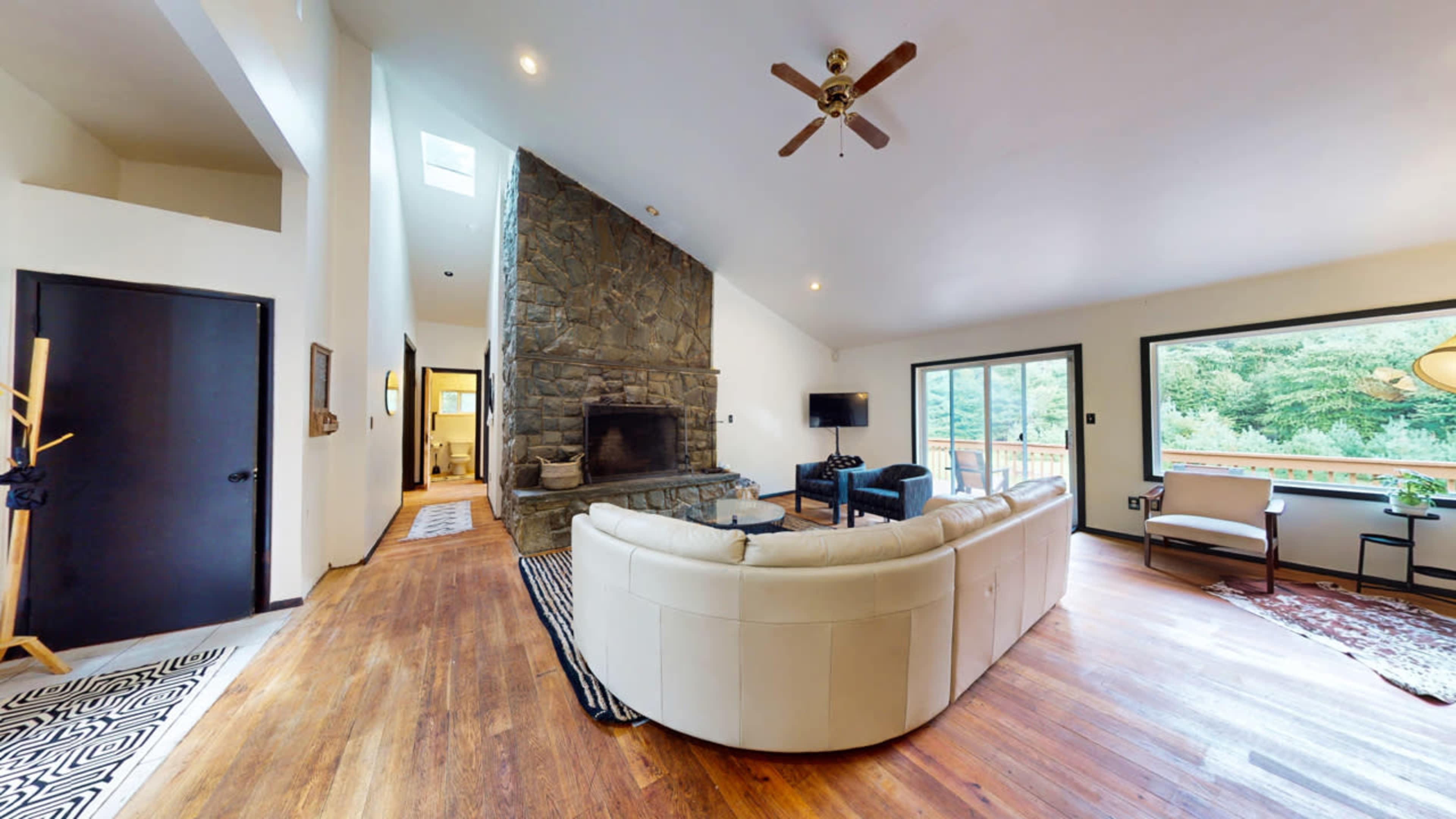 A spacious living room featuring a large stone fireplace, a curved sectional sofa, and windows that provide a view of surrounding greenery.