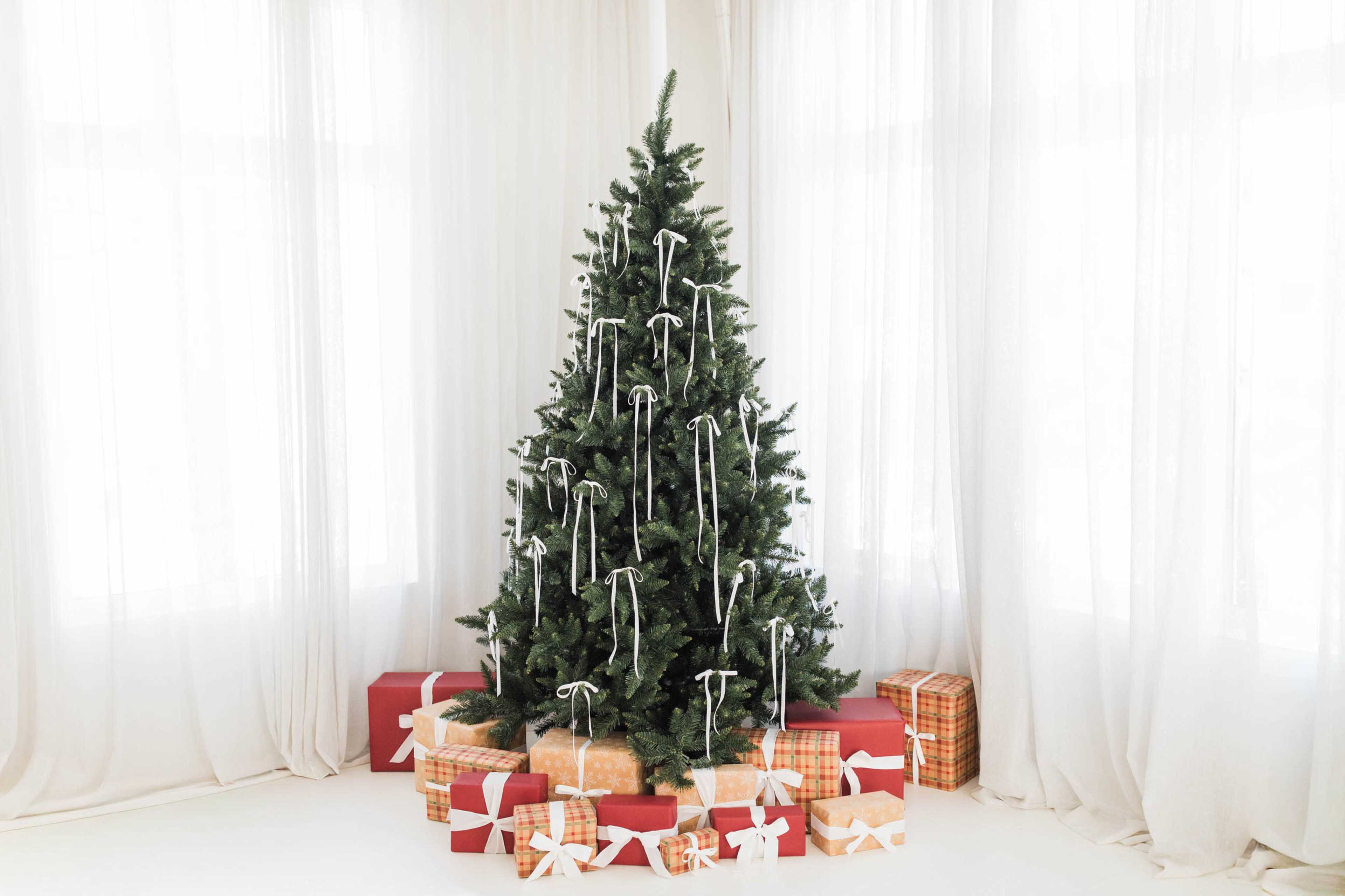 A Christmas tree adorned with ribbons stands among wrapped gifts in a bright, airy room.