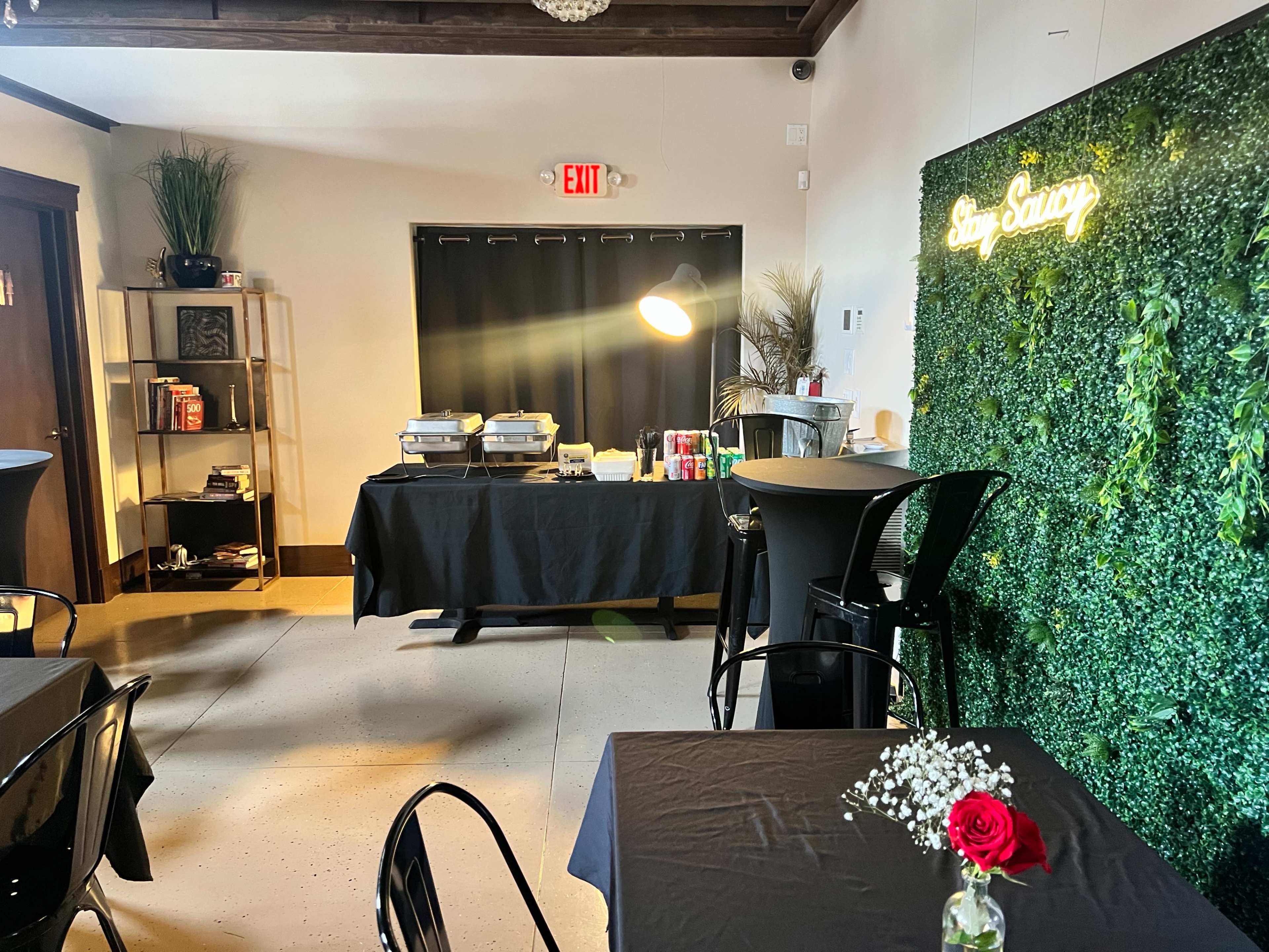 The image shows a decorated indoor event space with black tables, a food setup, and a green wall featuring a neon sign that reads "Stay Savory."