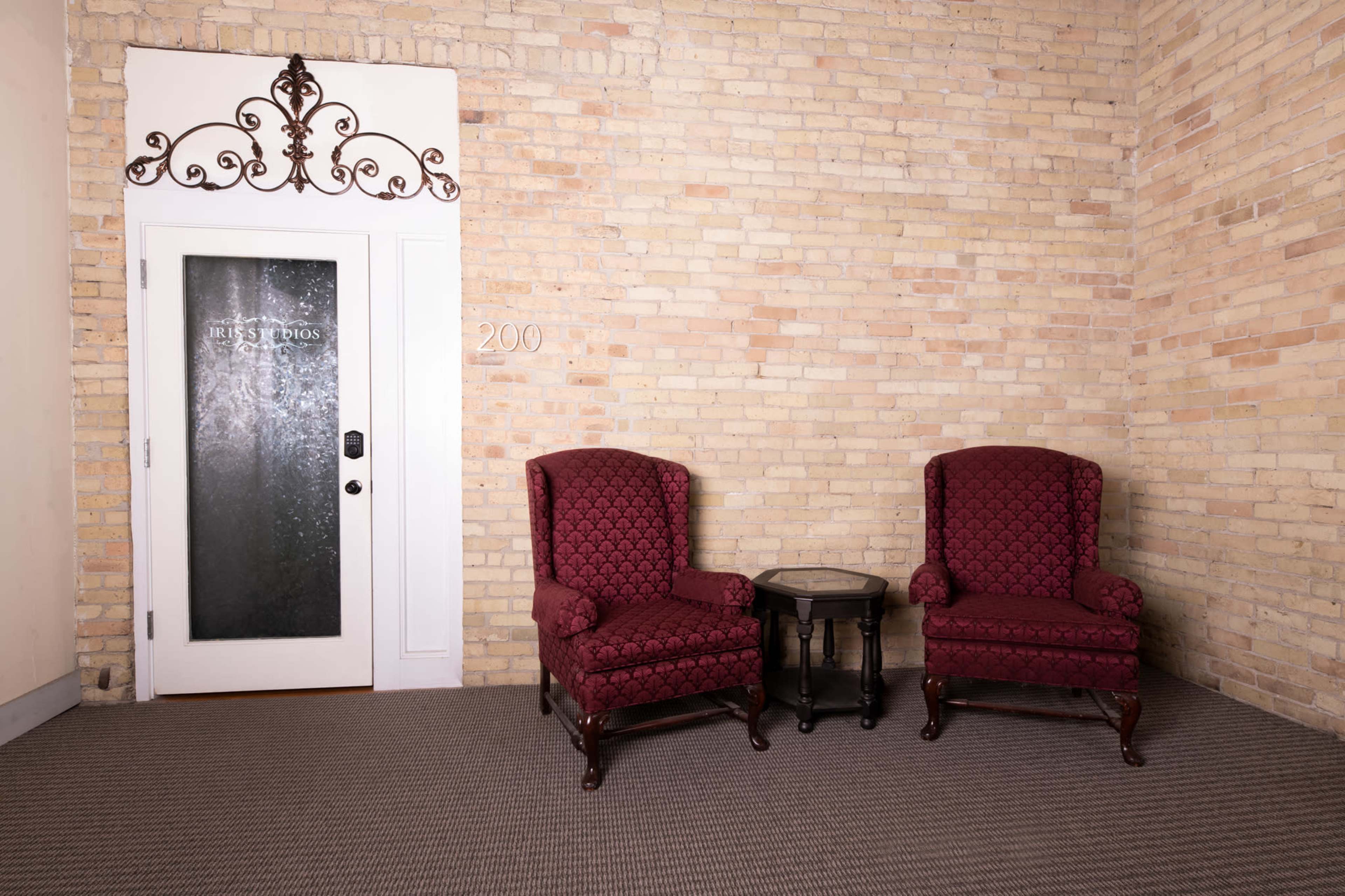 A corner of a room with two maroon upholstered chairs facing a small side table, next to a door with decorative elements and the number "200" on the wall.