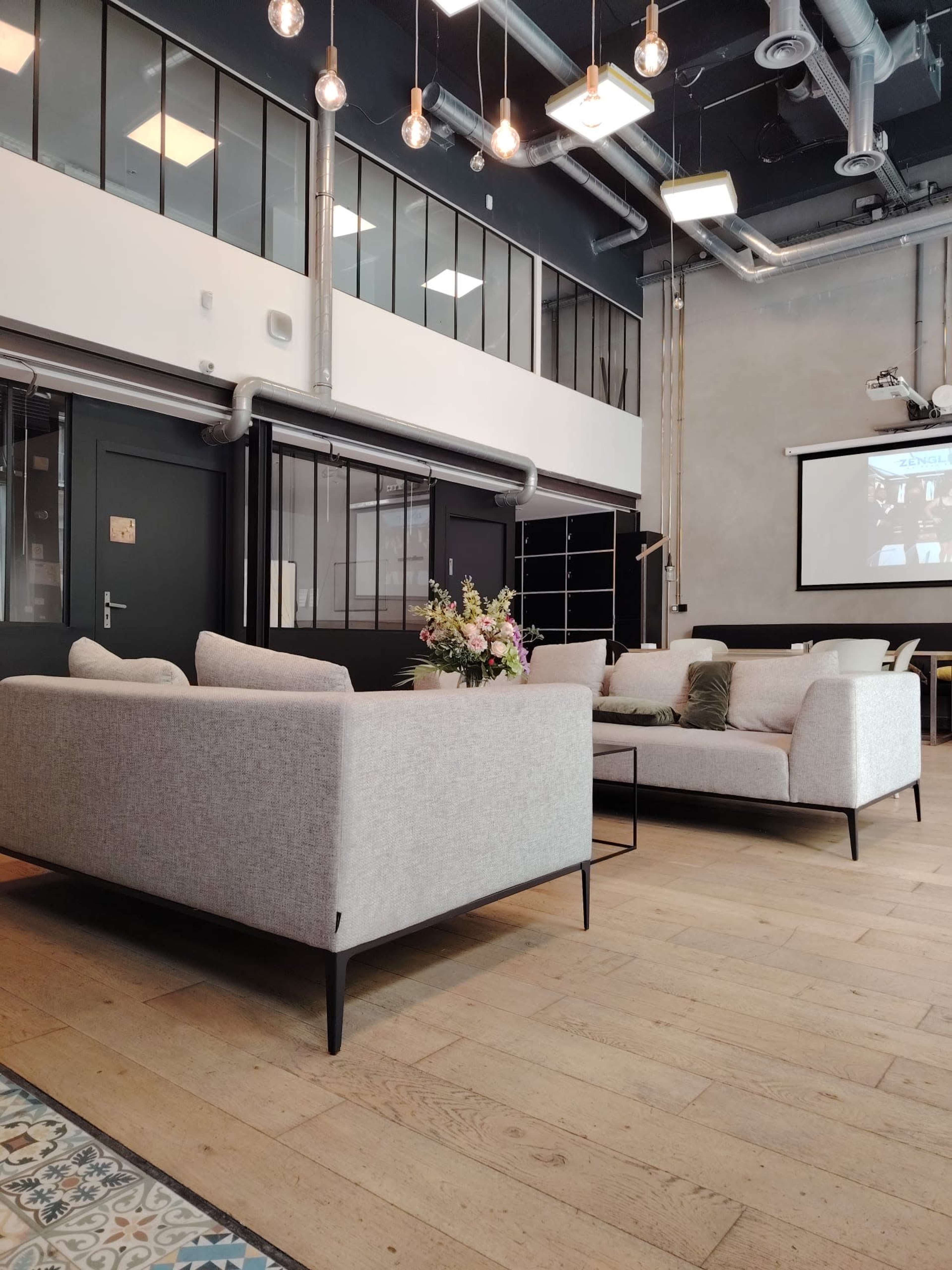 The image shows a modern lounge area with two light gray sofas, wooden flooring, and large windows allowing natural light, complemented by decorative lighting overhead.