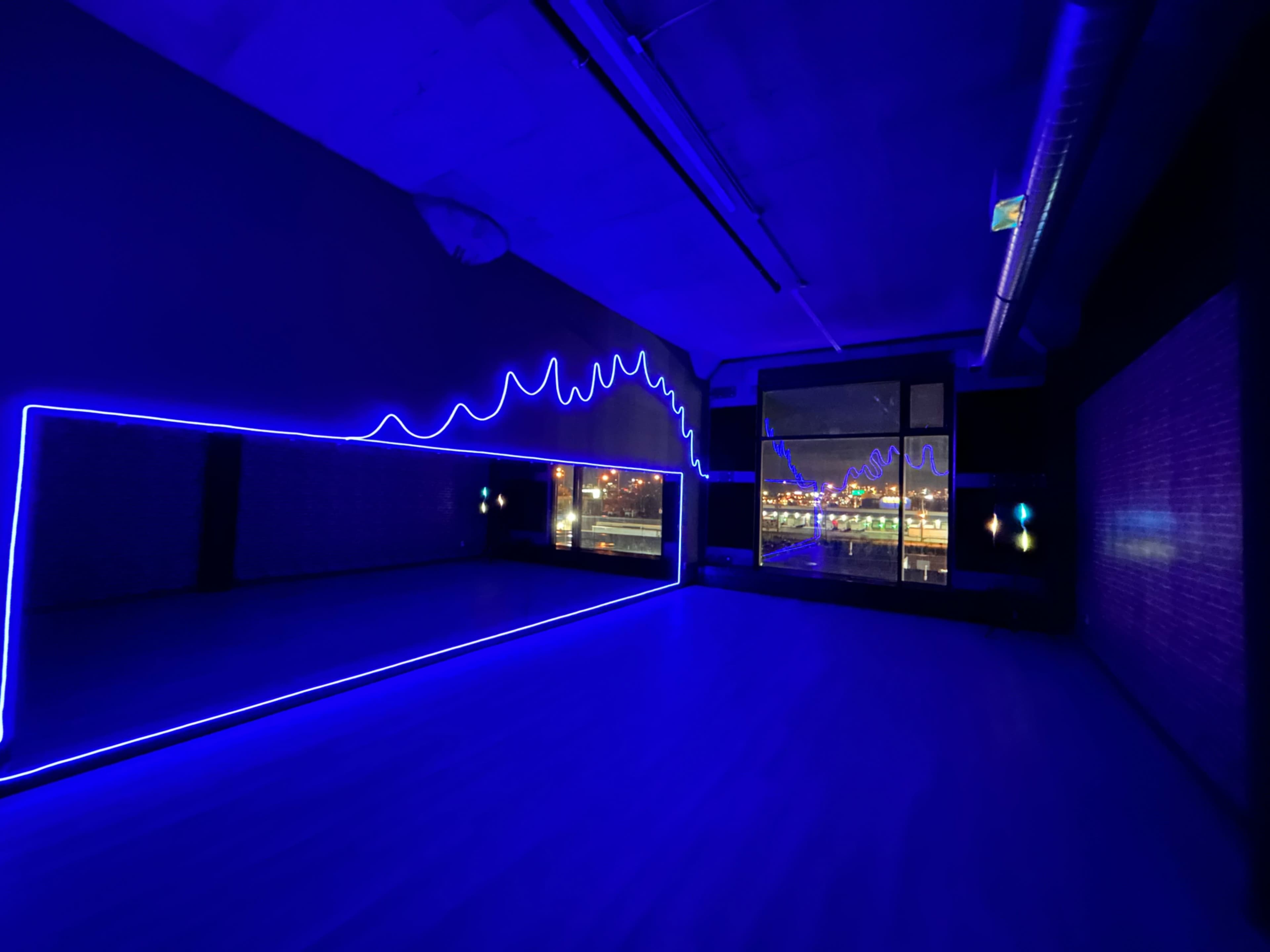 A dimly lit studio features a large mirror outlined with bright blue neon lights and a window displaying a city skyline at night.
