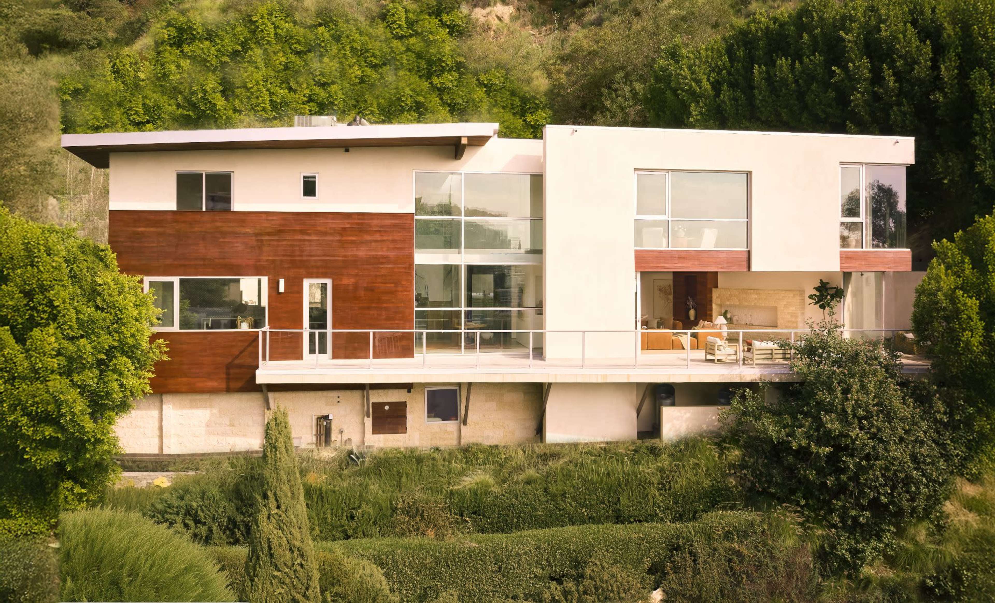 The image shows a modern two-story house with a mix of wood and white exterior, situated on a sloped landscape surrounded by greenery.