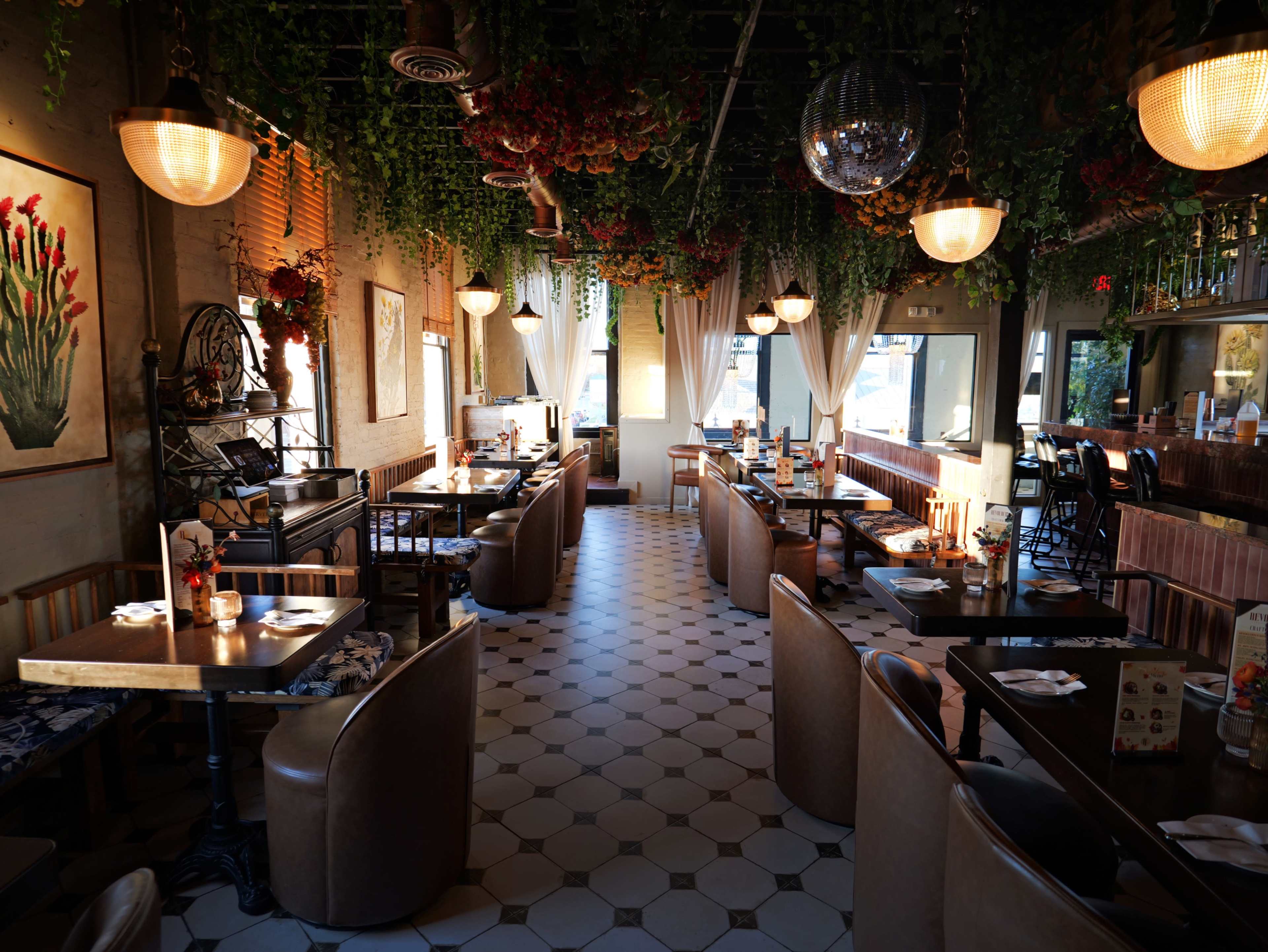 The image shows a restaurant interior featuring hexagonal tiled flooring, wooden tables with leather seating, and overhead light fixtures surrounded by hanging greenery and floral decorations.